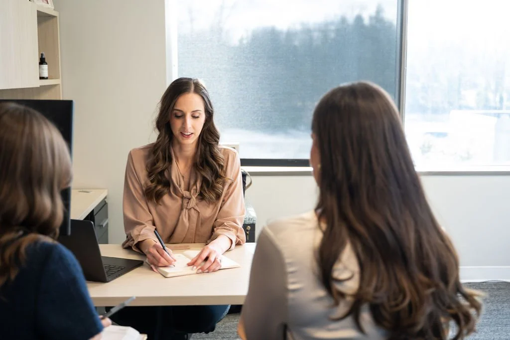 Three women having a meeting in an office, with one woman taking notes and the other two listening.