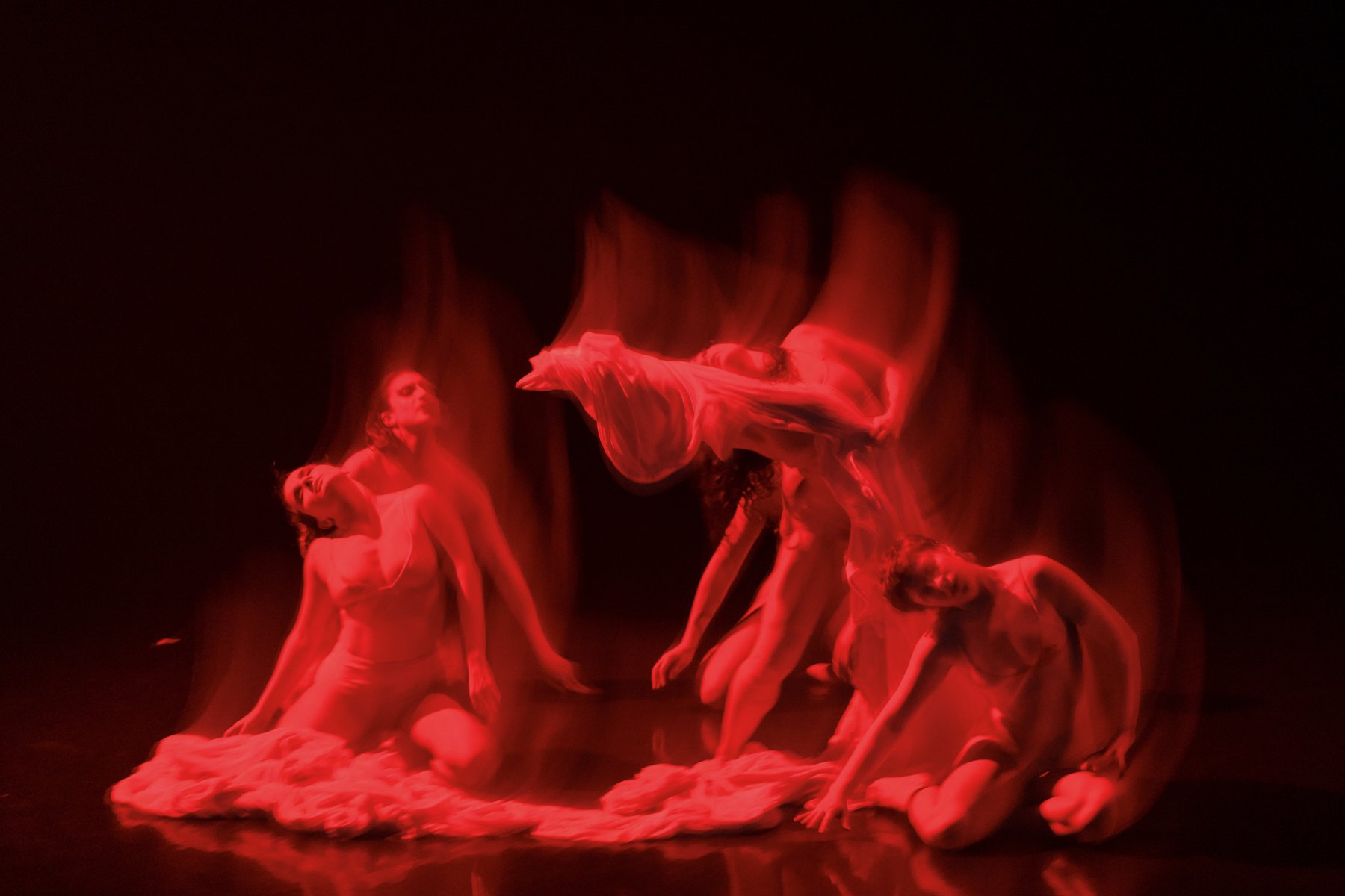 A dance performance with multiple women dressed in flowing white costumes, illuminated by red lighting, on stage with dramatic and artistic poses.