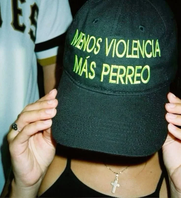 A person is holding a black baseball cap with green embroidery that says 'Menos Violencia Más Perreo'. The person is wearing a black tank top, a cross necklace, and a ring on the finger of their left hand.