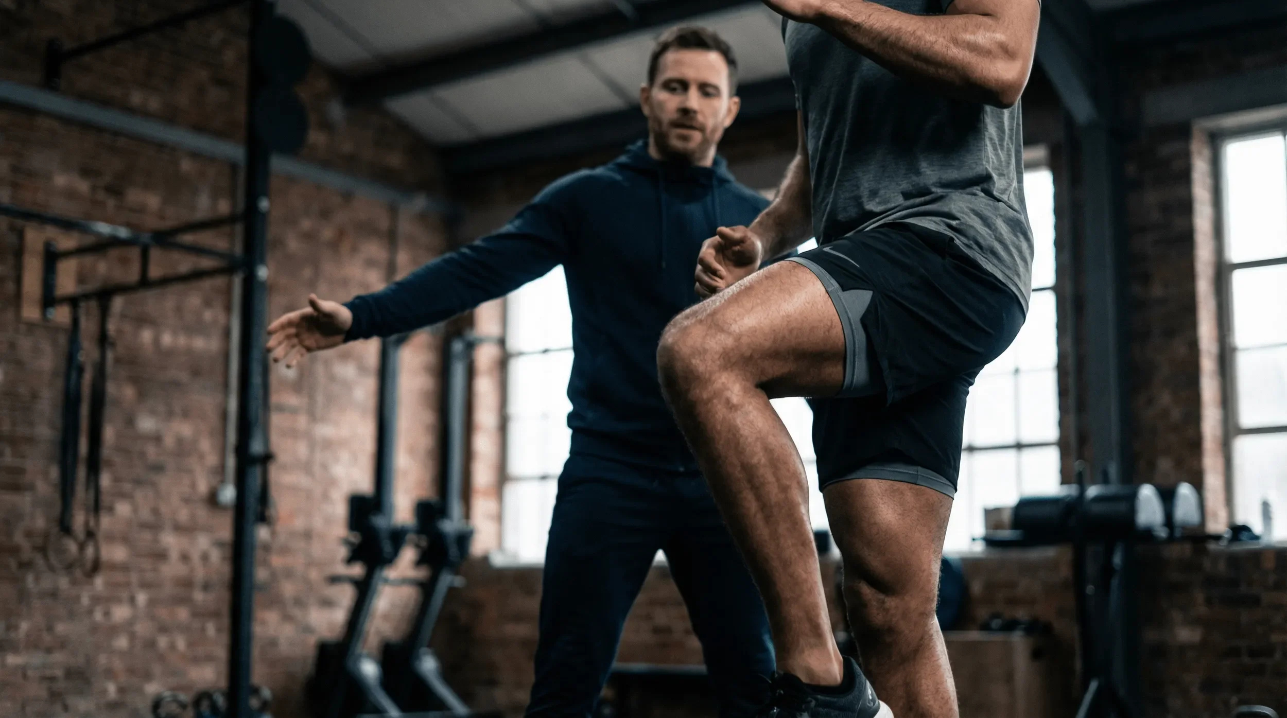 A personal trainer is instructing a man during a workout session in a gym. The man is performing a knee lift exercise while the trainer demonstrates with his arms outstretched for guidance. The gym has brick walls and large windows.