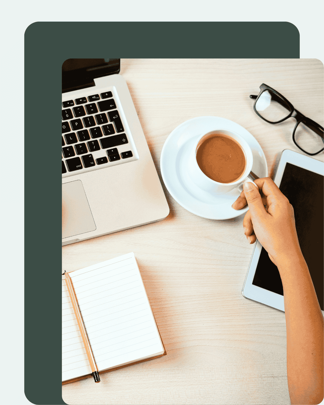 Top-down view of a workspace with a laptop, a cup of coffee on a saucer, a pair of glasses, a tablet, an open lined notebook, and a person's hand holding a spoon over the coffee.