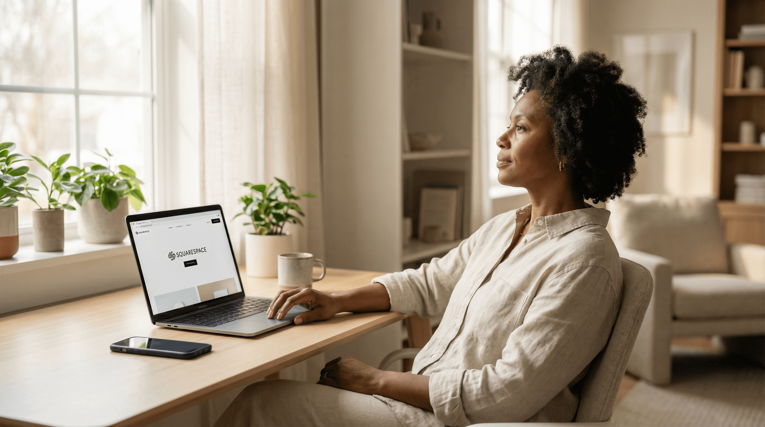 A woman sitting at a desk using a laptop with Squarespace website open, with plants and a window in the background