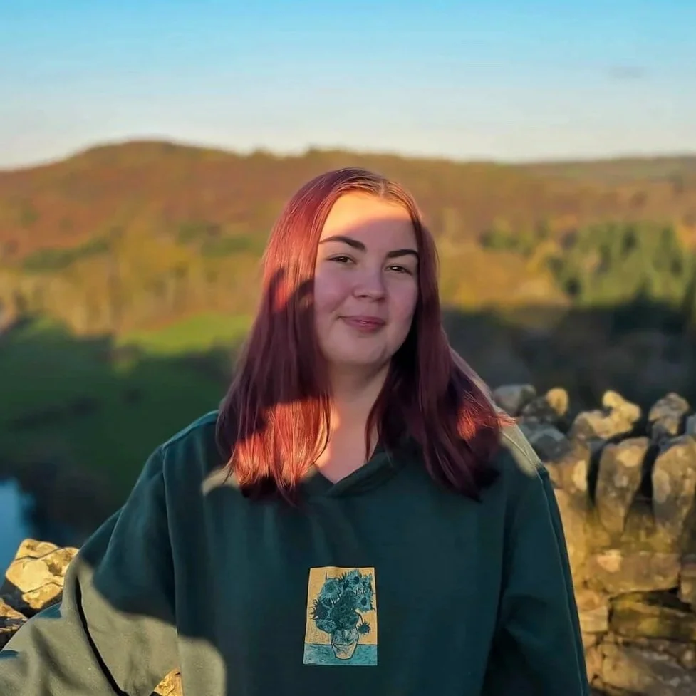 A young woman with reddish-brown hair standing outdoors near a rocky area, with green rolling hills and a body of water in the background during sunset.