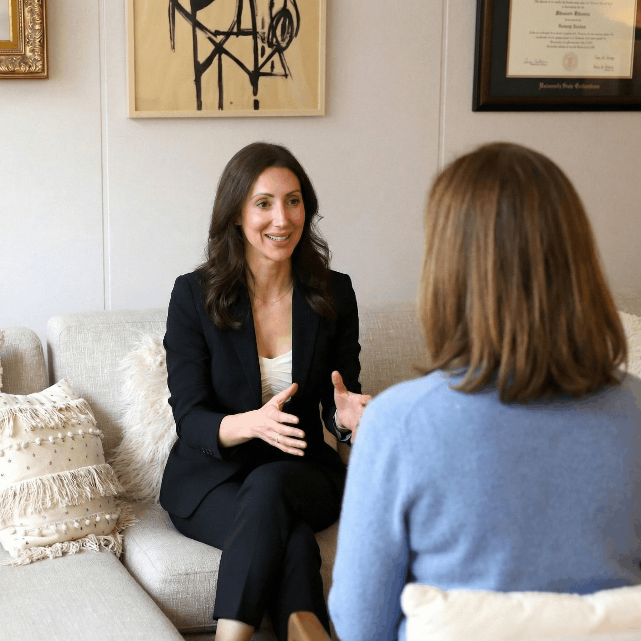 Two women are sitting on a beige sofa, engaging in a conversation in an office or therapy setting. One woman, dressed in a black blazer and white top, is gesturing with her hands and speaking, while the other, with shoulder-length brown hair and a blue sweater, listens attentively. The background has framed artwork and a diploma on the wall.