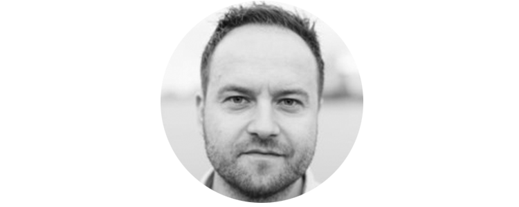 Black and white close-up of a man with short hair and slight beard, looking directly at the camera.