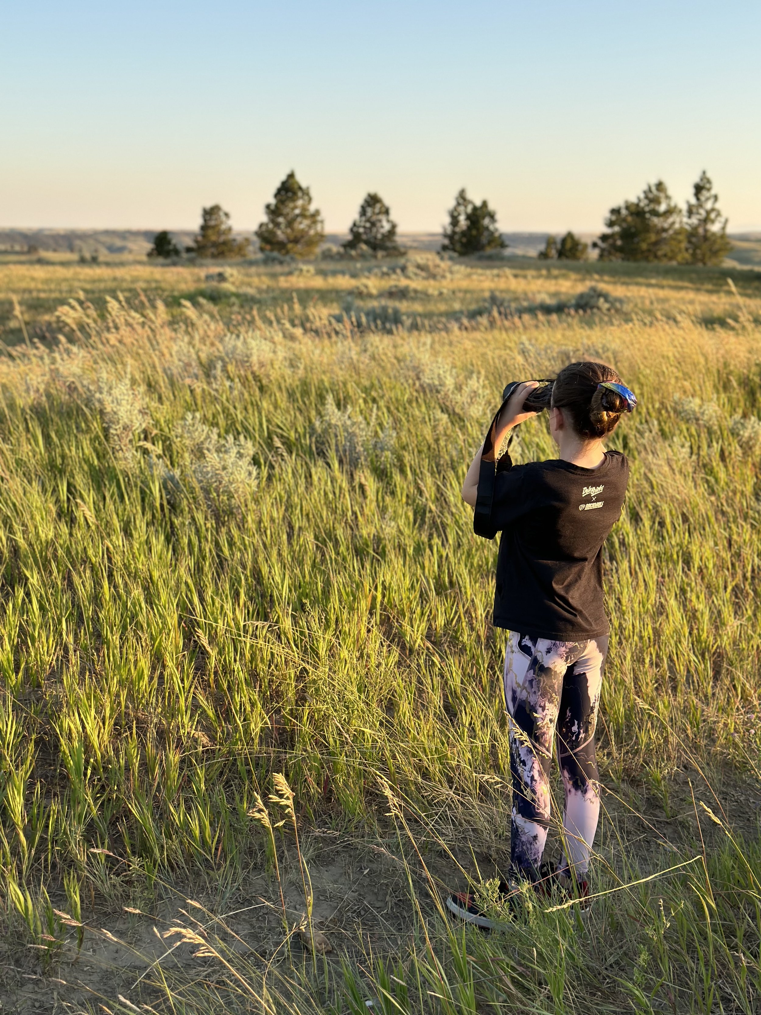 A woman standing in a grassy field taking a photo with a camera during sunset, with trees in the background.