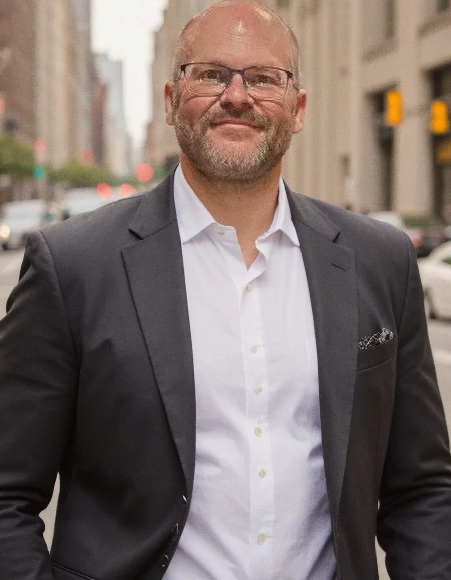 A middle-aged man with glasses, a beard, and a bald head, wearing a navy blue suit, white shirt, and dark tie, smiling while walking outdoors in an urban area with tall buildings and people in the background.
