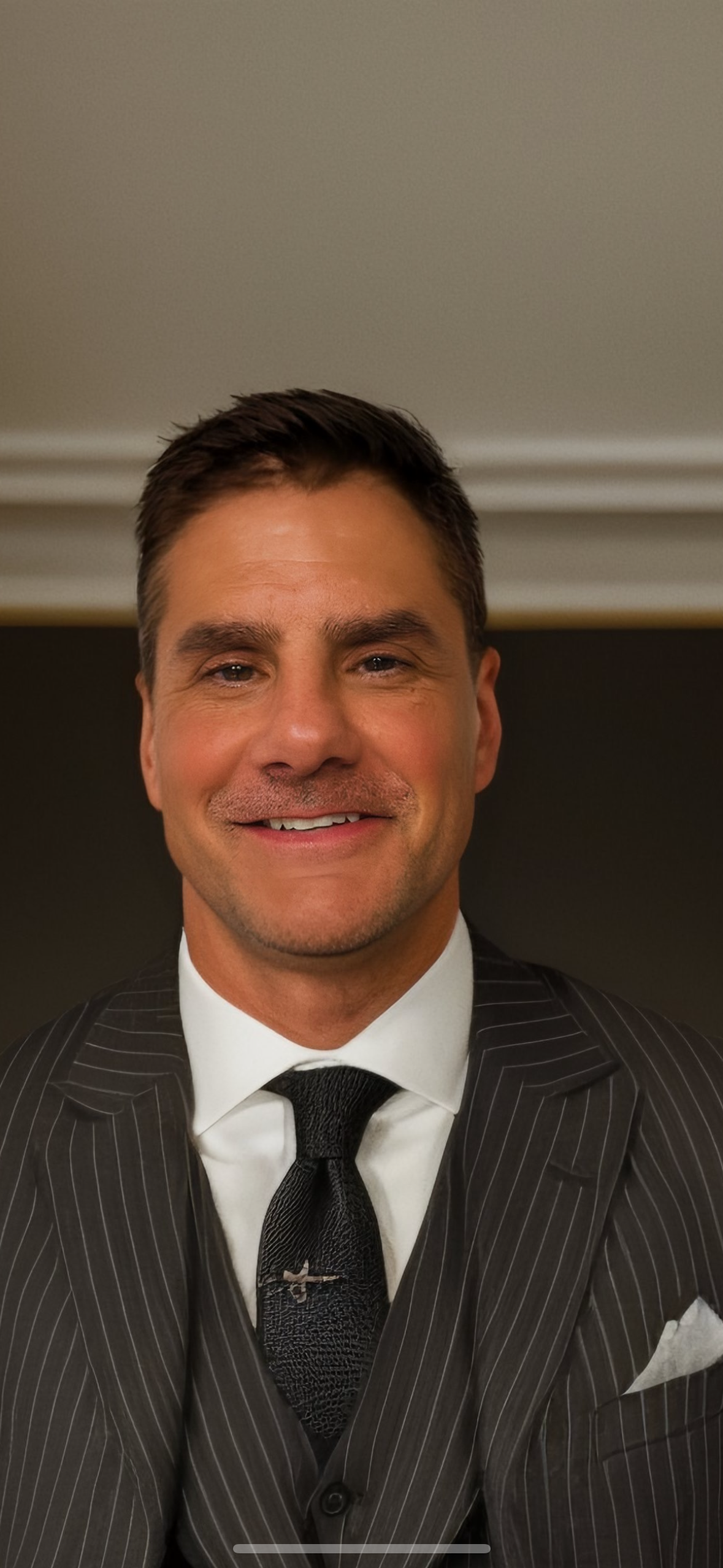 A man wearing a black pinstripe suit, white shirt, and black tie, smiling at the camera in an indoor setting.