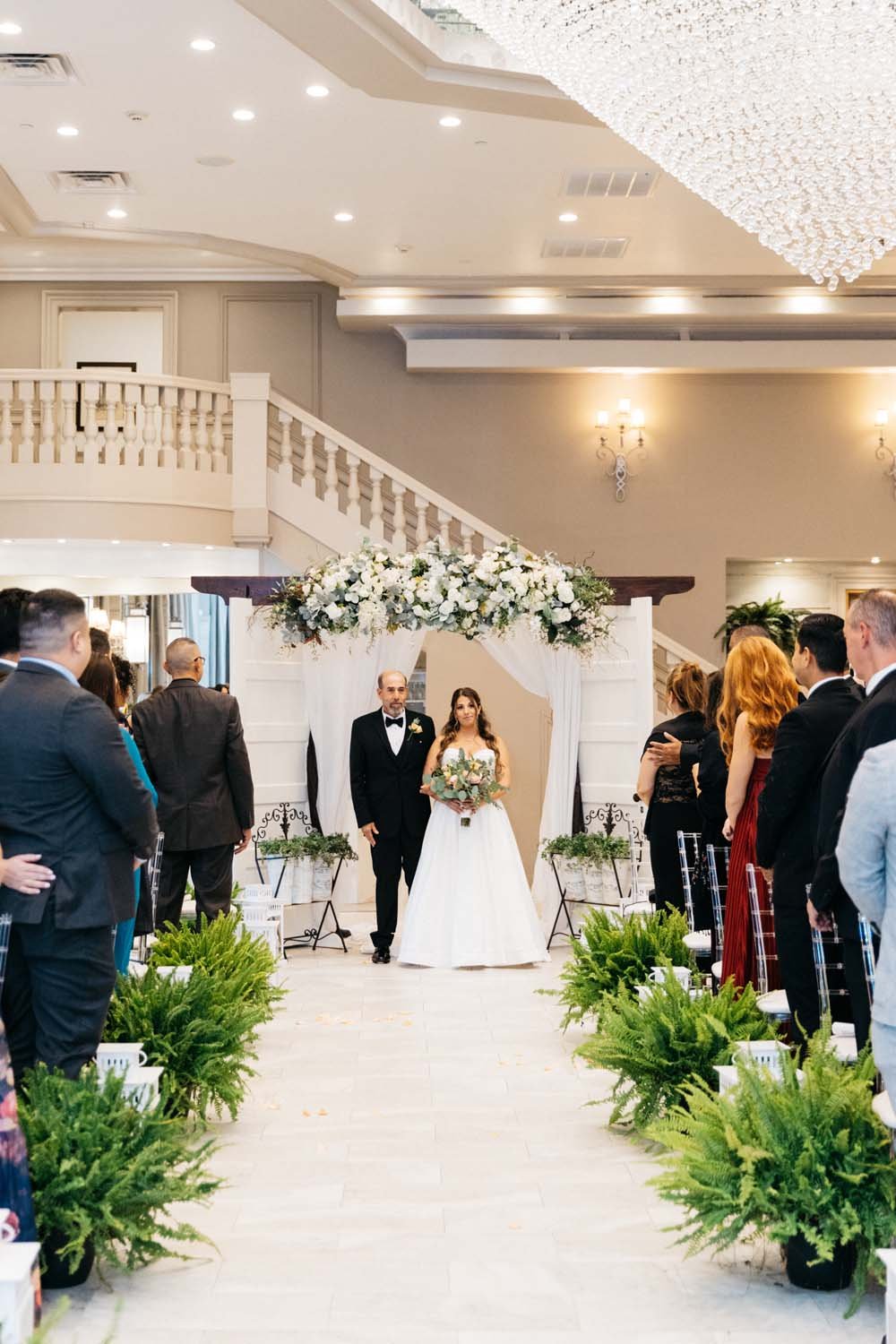 A bride and her father walking under a magnificent white floral arbor at the back of a luxury ballroom venue in upstate NY