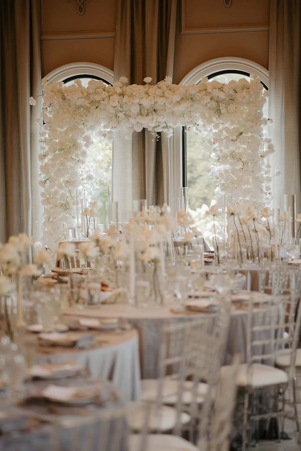 View of a grand ballroom filled with silver chiavari chairs through an all white floral arch