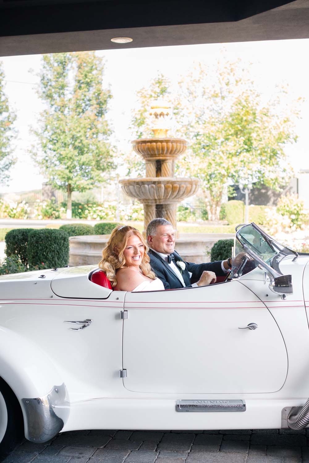 Bride and her father smiling inside a classic vintage white convertible in front of the stone fountain at Villa Venezia.