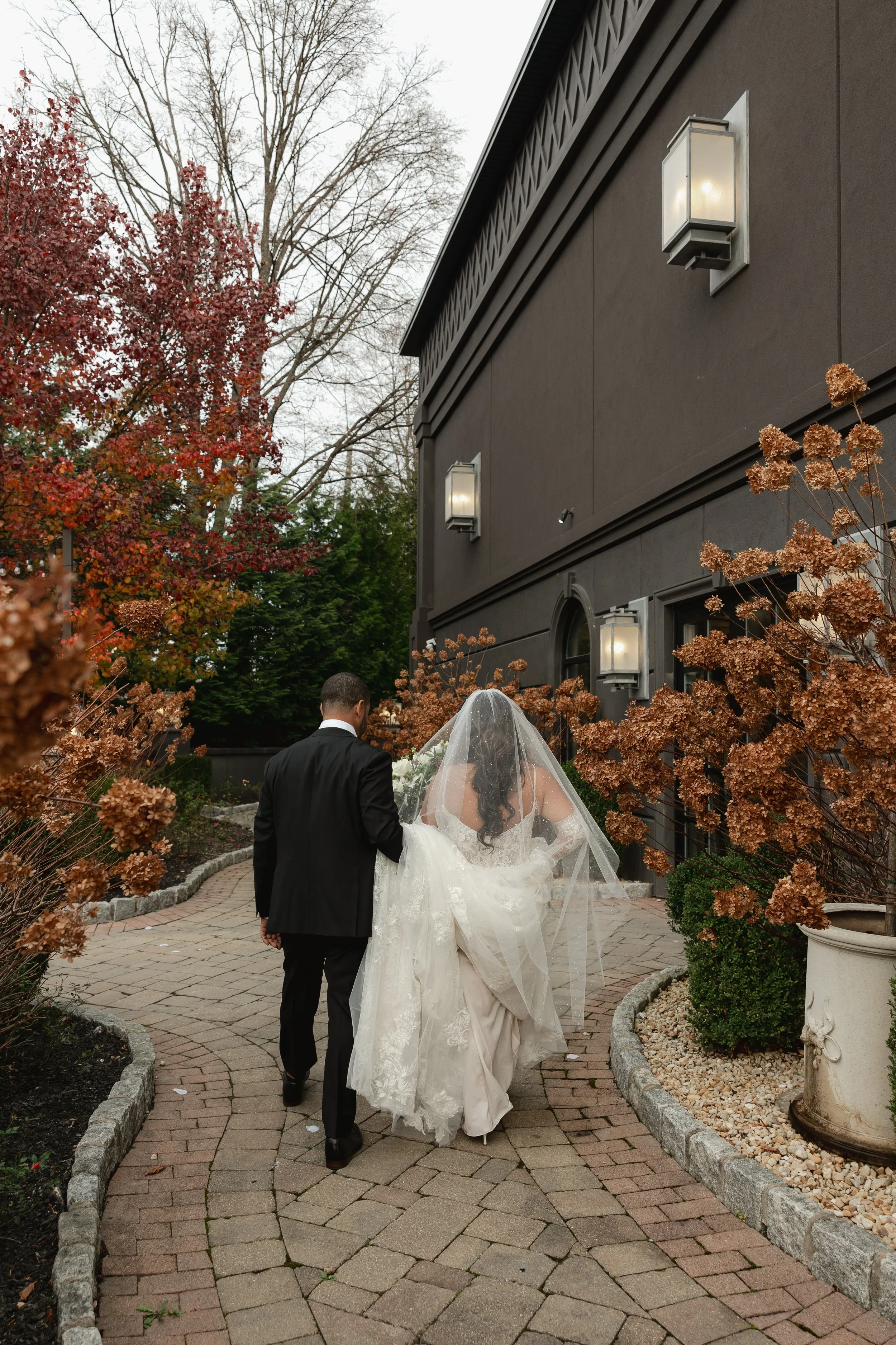 A bride and groom walk through the manicured gardens at Villa Venzia after the leave have changed in Fall, featuring rich reds and warm browns.