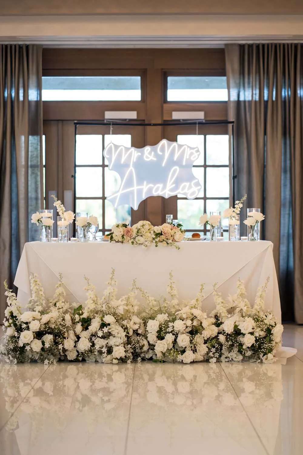 A luxurious wedding sweetheart table featuring lush white floral runners, floating candles, and a custom neon name sign.