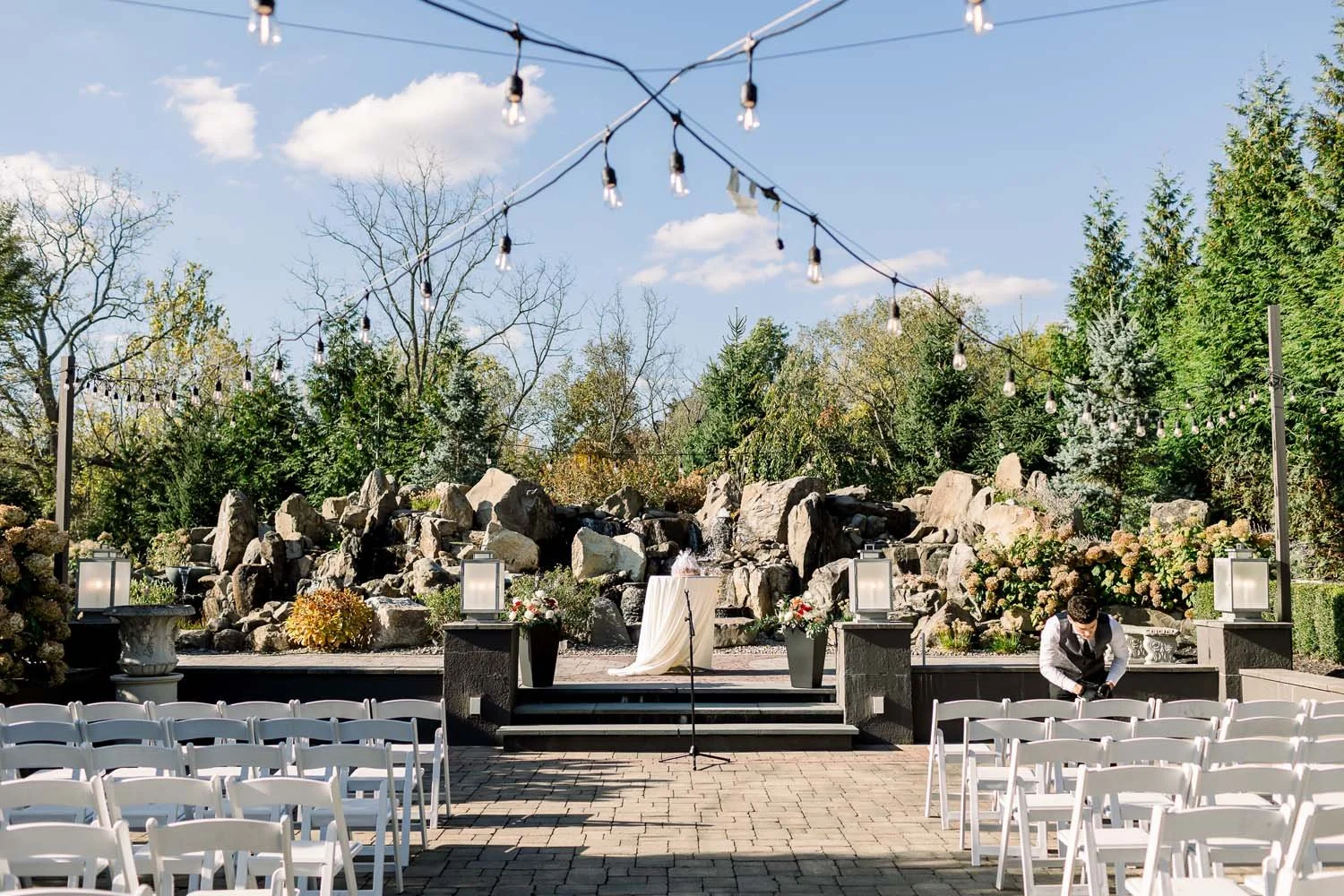 Wide shot of an outdoor Hudson Valley wedding venue courtyard with white chairs, string bistro lights, and a stone waterfall altar at Villa Venezia