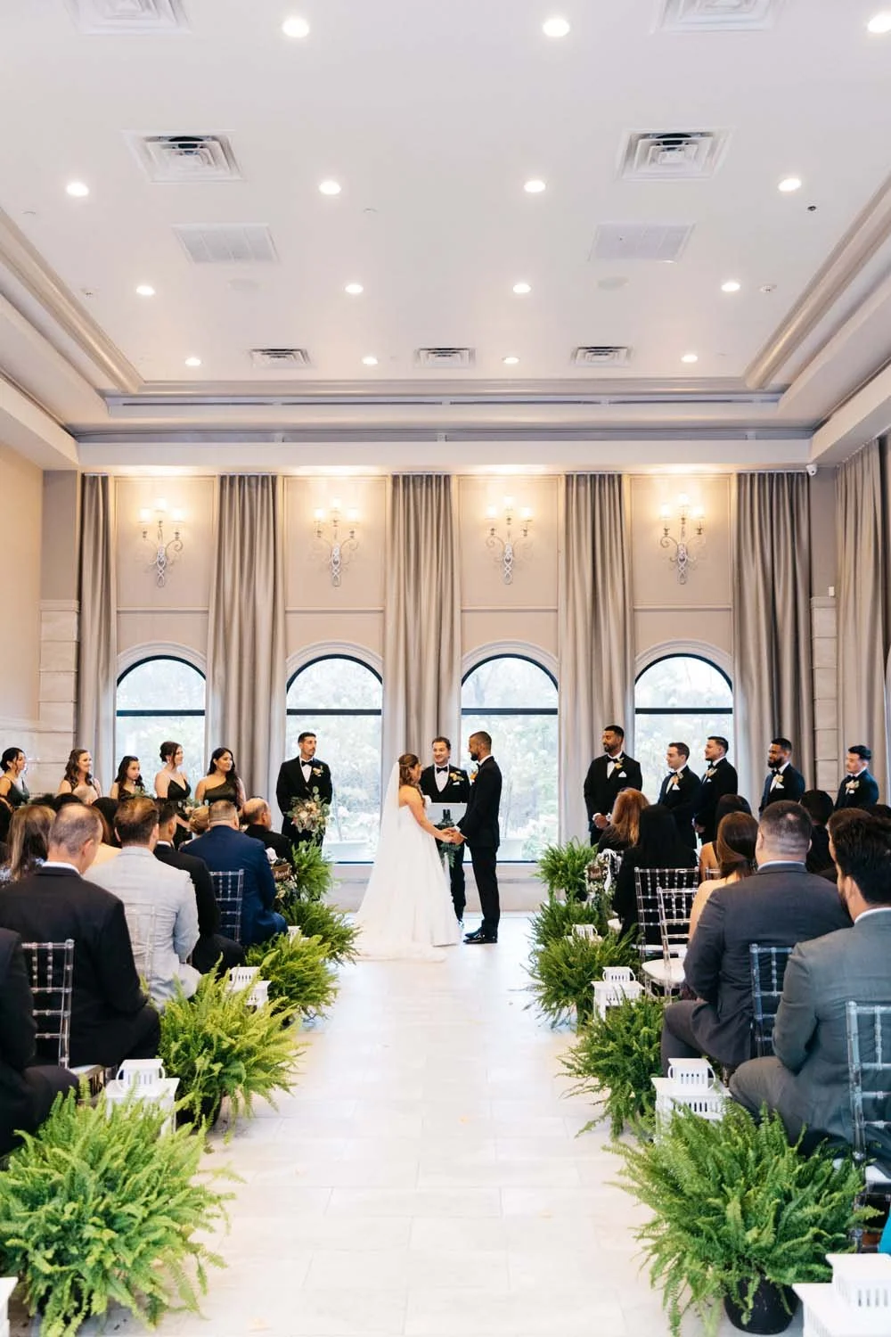 Grand ballroom wedding ceremony setup with large arched windows and lush greenery lining the aisle at Villa Venezia