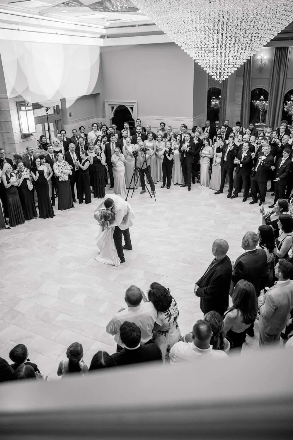 Black and white overhead shot of a bride and groom dipping during their first dance under a massive crystal chandelier in the Villa Venezia ballroom.