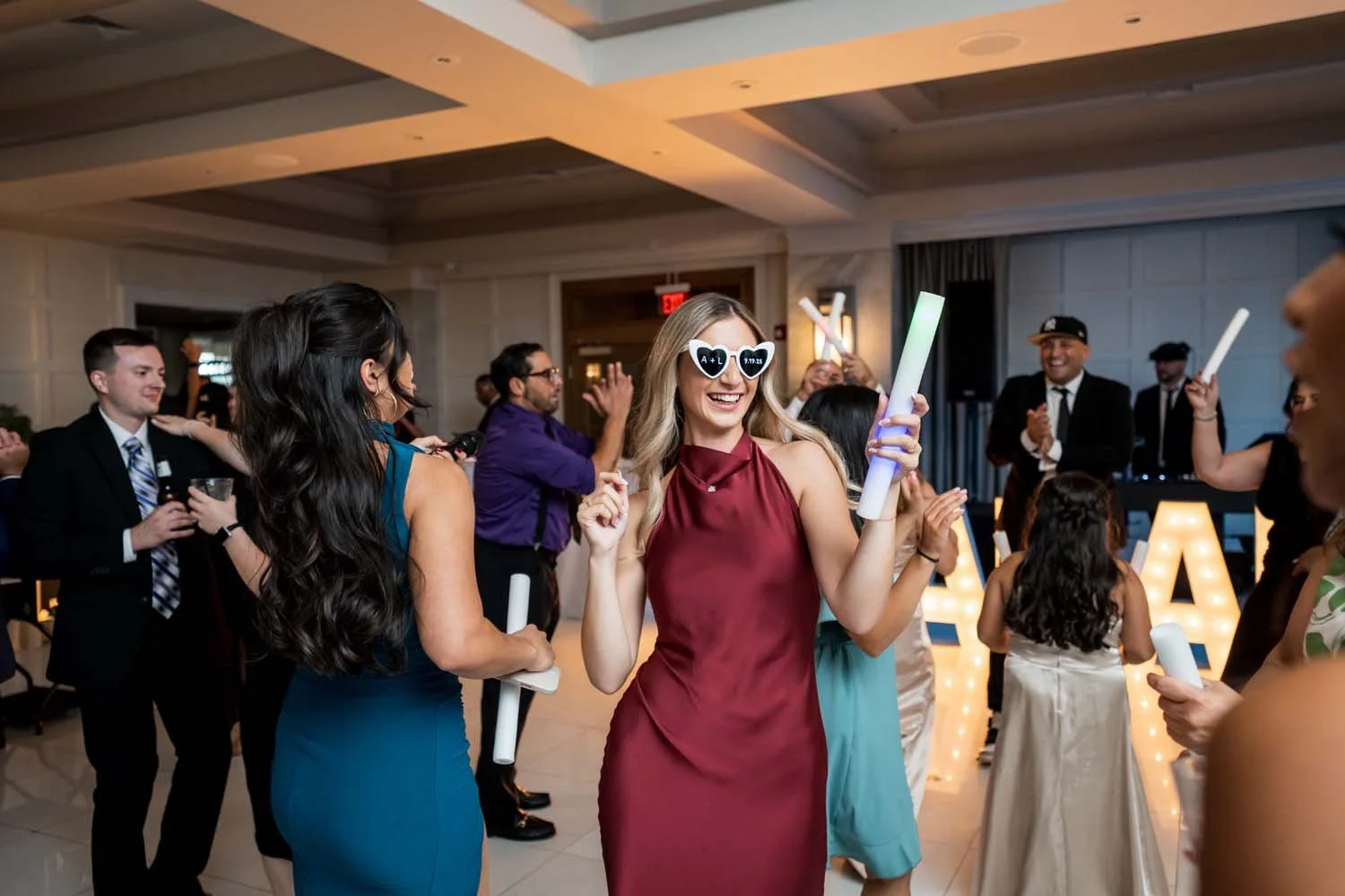 Wedding guests laughing and dancing with white foam glow sticks on a spacious ballroom dance floor.