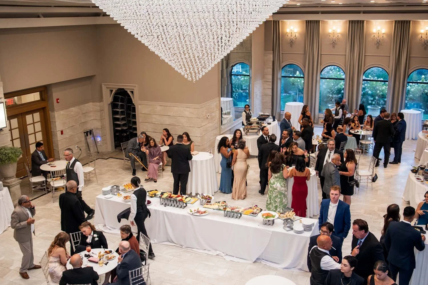 Overhead bird's-eye view of wedding guests mingling around food stations during a cocktail hour beneath a giant crystal chandelier.