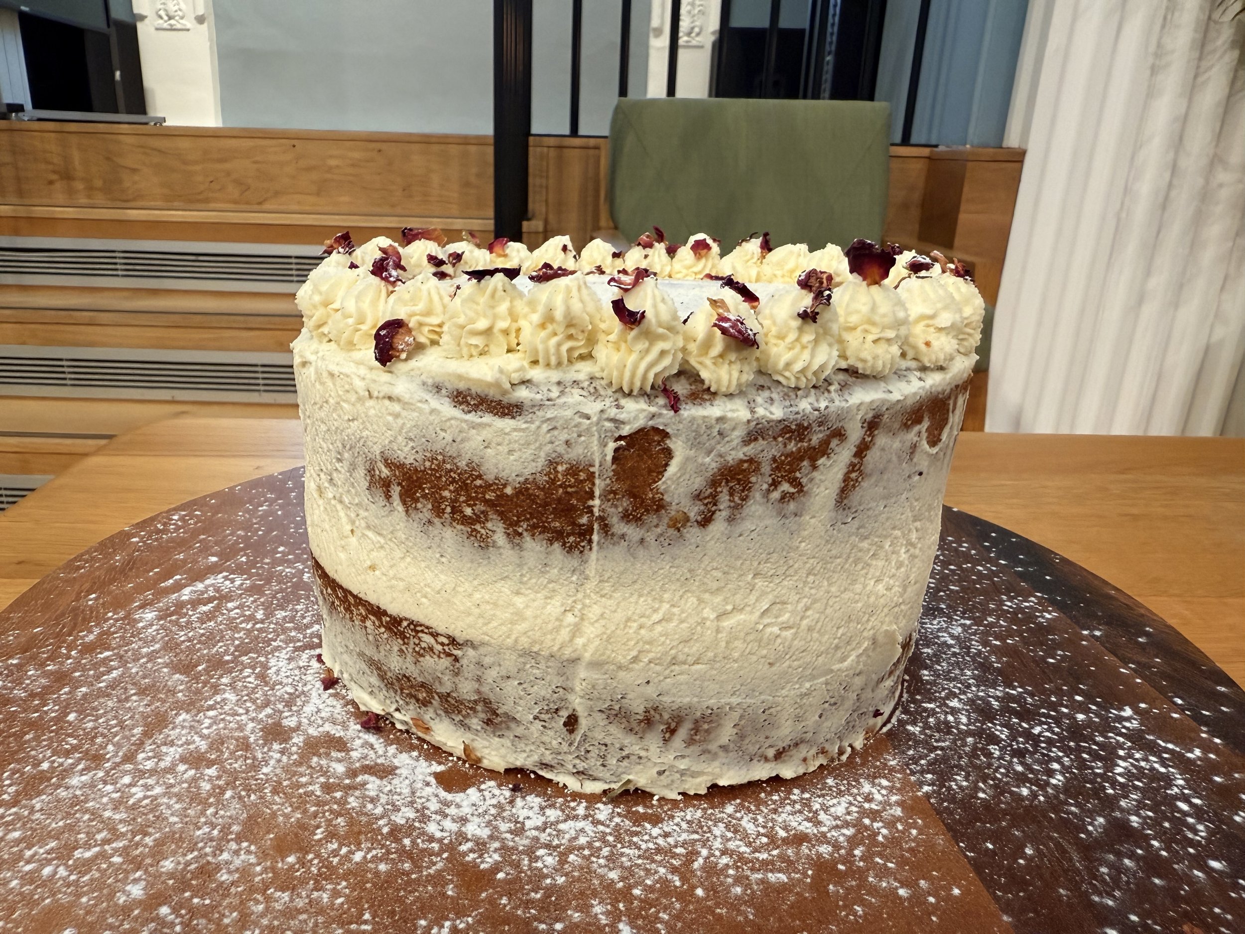 A tall layered vanilla cake with frosting and cream on top, decorated with small piped frosting rosettes and dried flower petals, placed on a wooden surface dusted with powdered sugar, in a cozy indoor setting.
