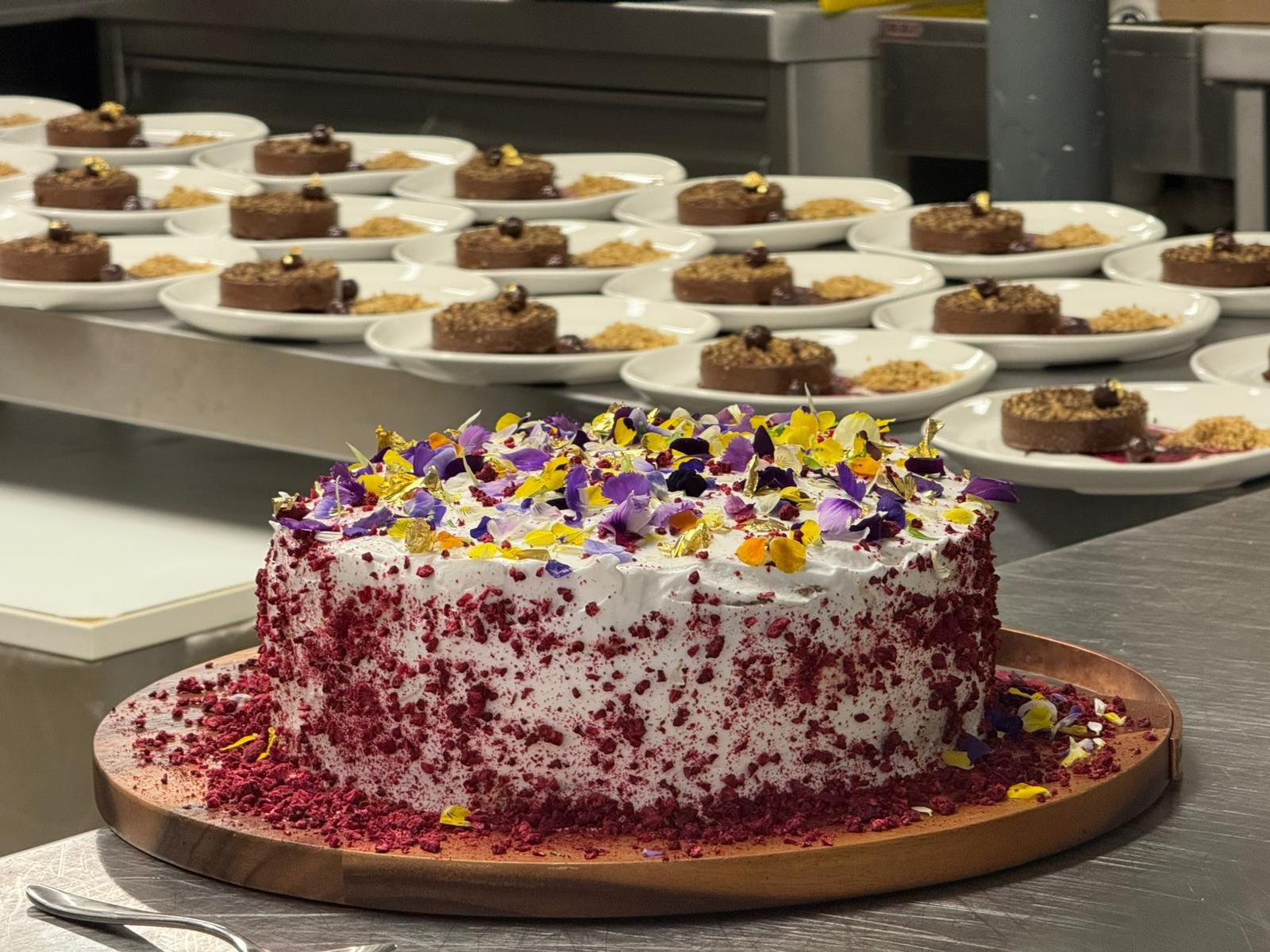 A decorated cake with white frosting, red crumbs, and colorful edible flowers in front of numerous plated desserts on a stainless steel countertop.