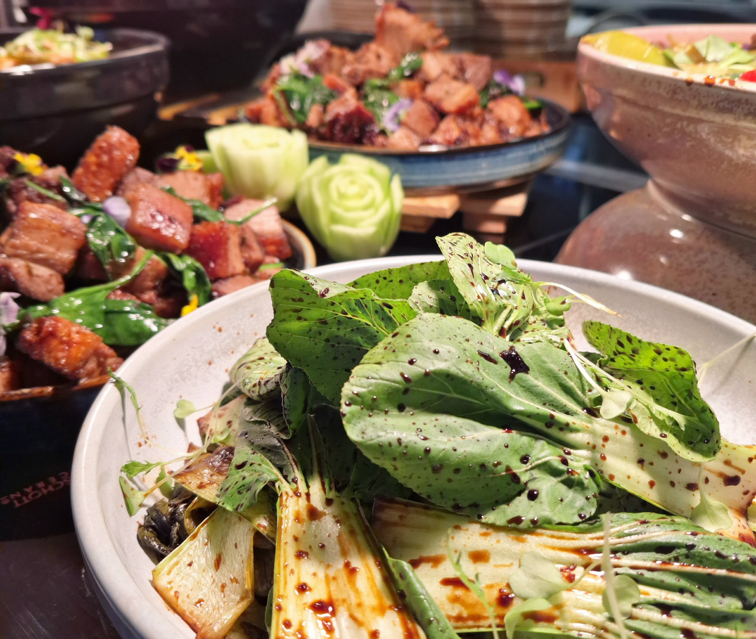 Close-up of a salad with mixed greens and dark dressing in a white bowl, with various plated dishes of grilled meats and vegetables in the background.