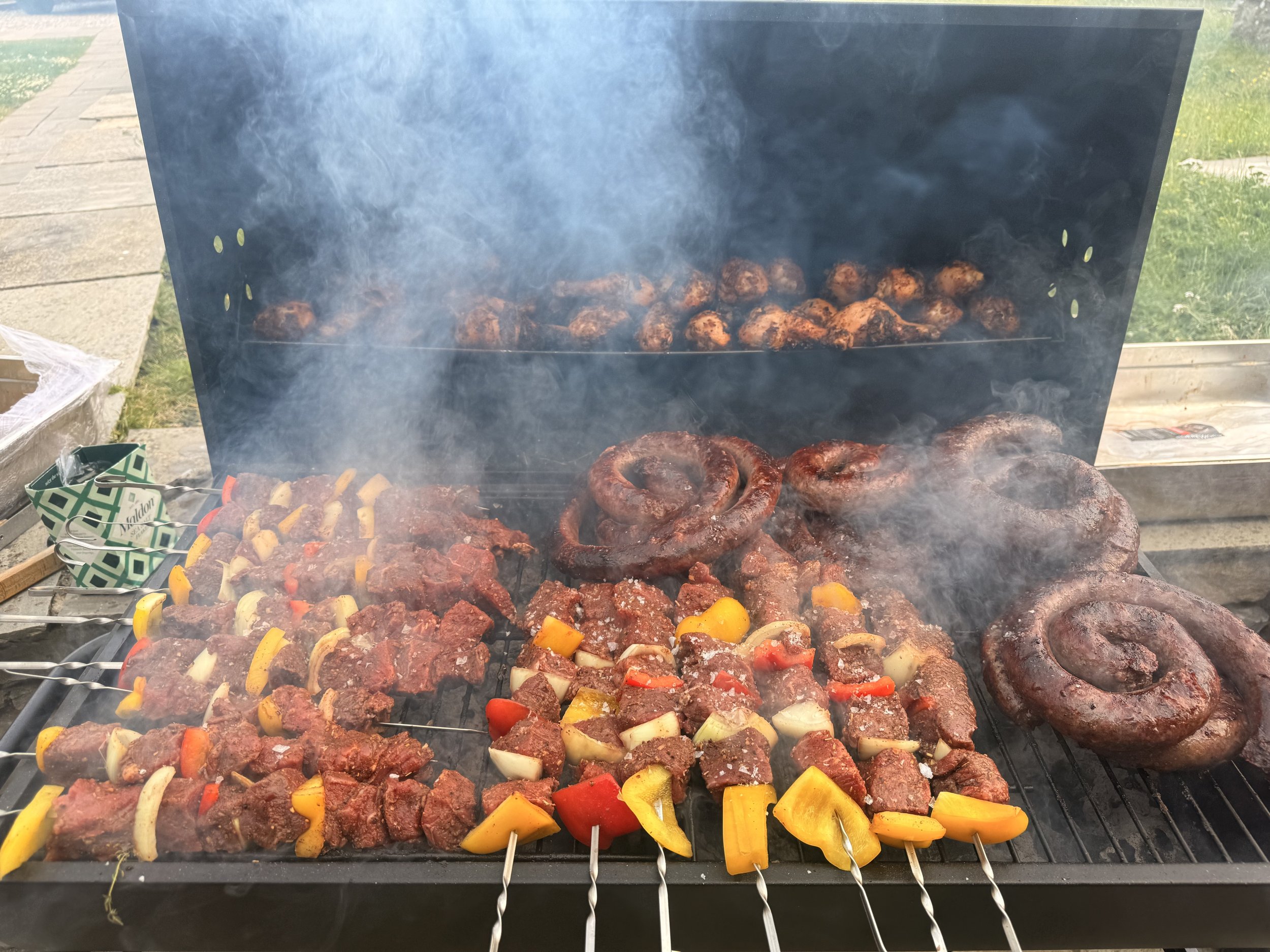 Barbecue grill with skewers of seasoned beef, chunks of vegetables, and large sausage coils, with smoke rising from the grilling food.