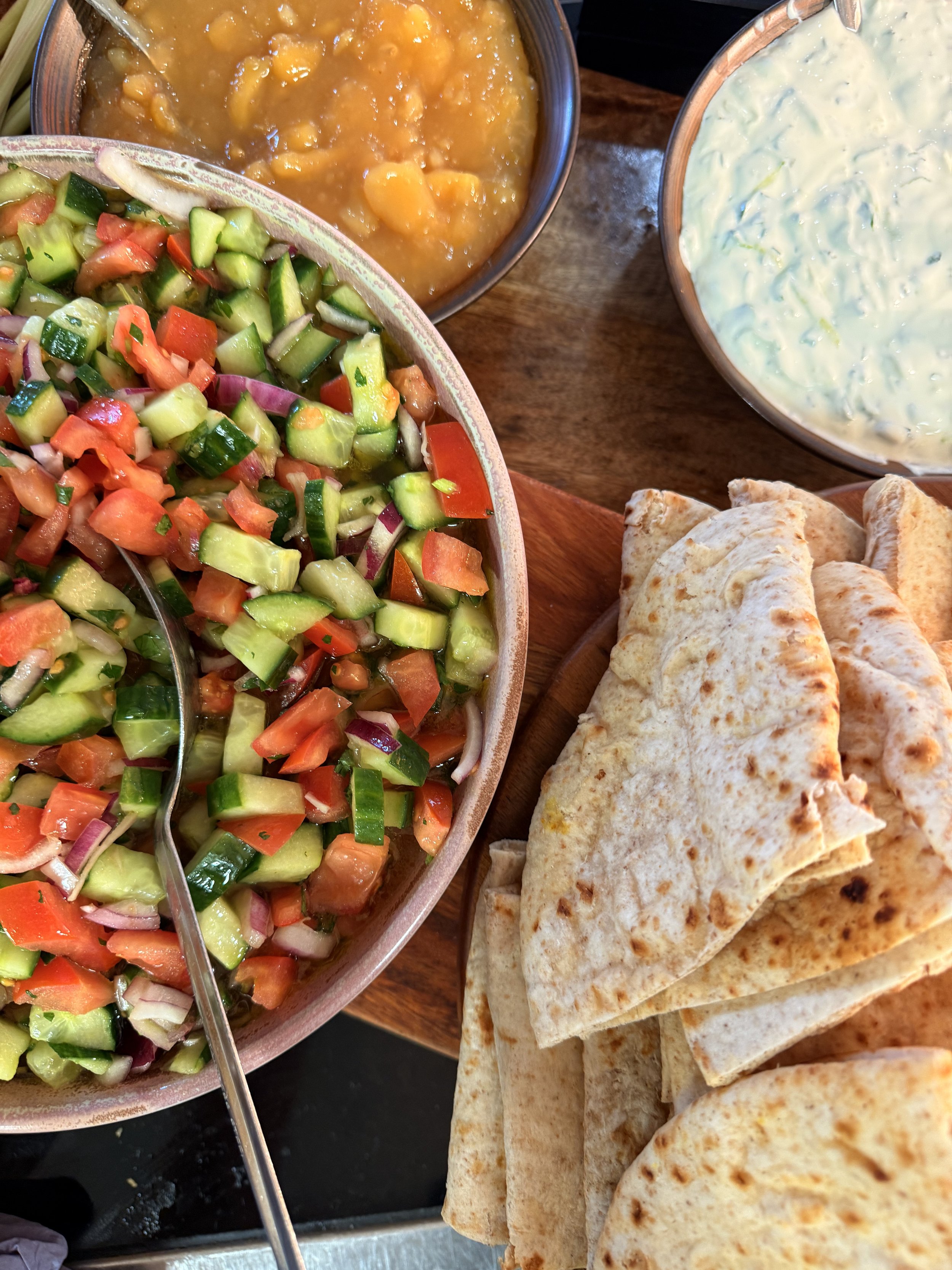 A platter of fresh chopped cucumber, tomato, and onion salad, alongside warm folded flatbreads and small bowls of yellow lentil stew, creamy cucumber sauce, and tangy chutney or salsa.