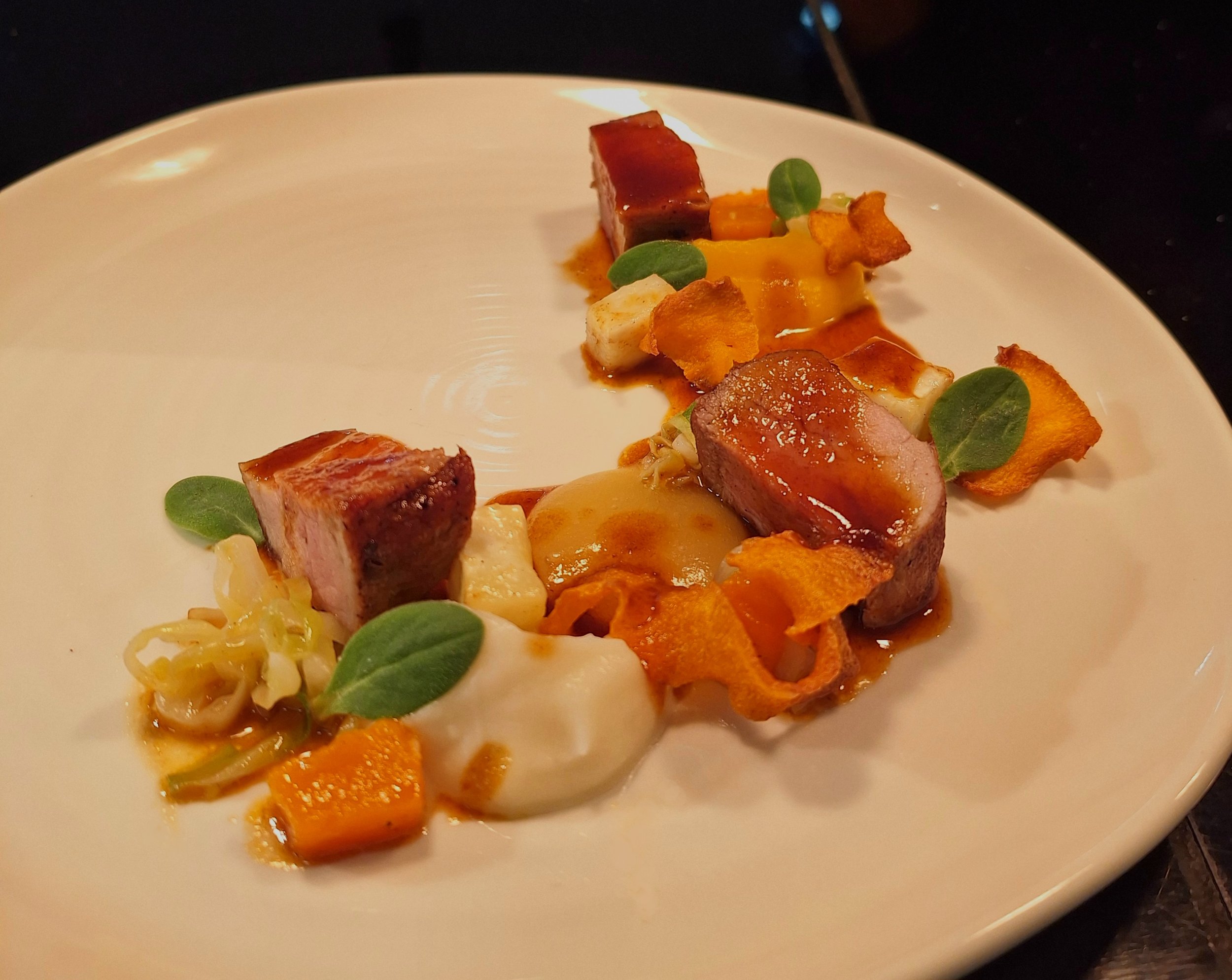 Fine dining dish with seared meat pieces, microgreens, roasted vegetables, herbs, and sauce on a white plate.