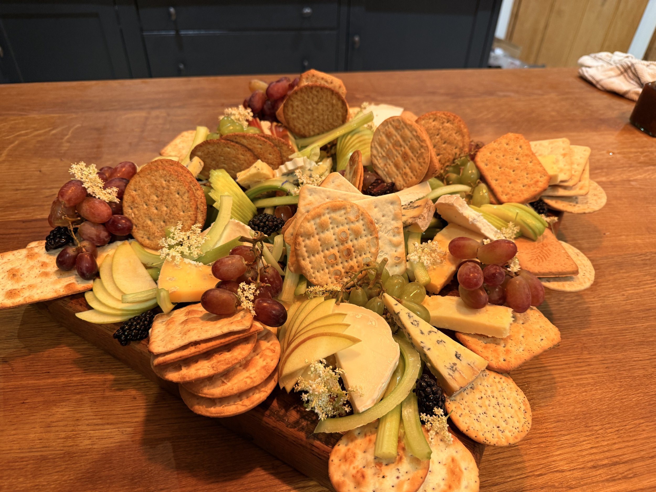 A cheese and cracker platter with grapes, celery, and flowers on a wooden table.
