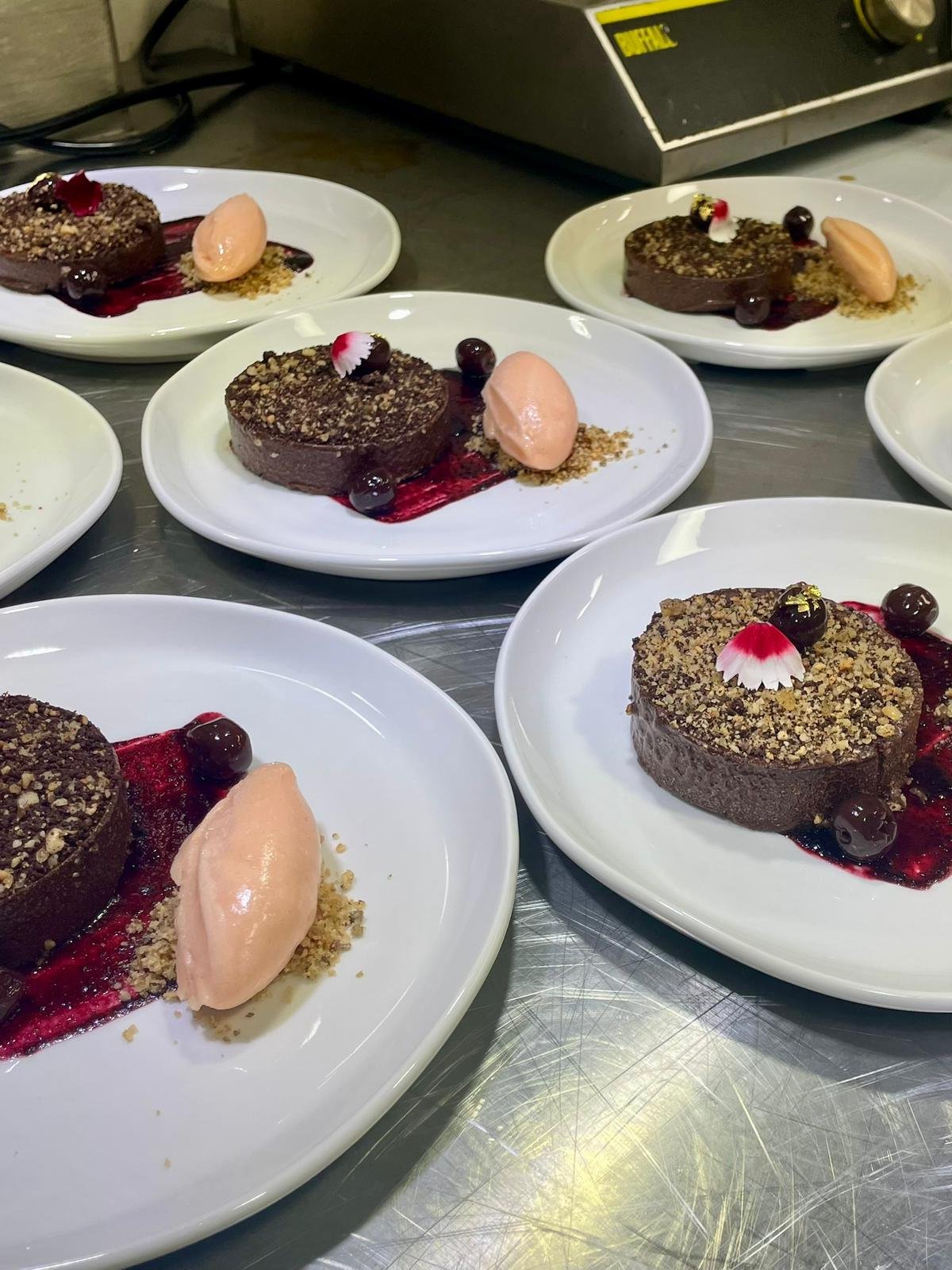 Six white plates with small chocolate desserts topped with berries and garnishes, arranged on a stainless steel table in a kitchen or food preparation area.