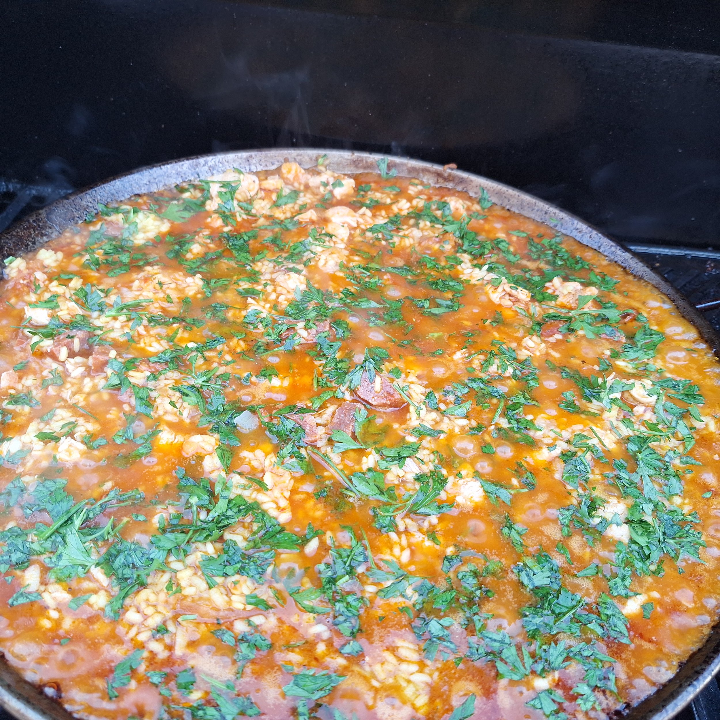 A close-up of a skillet containing a simmering dish with chopped herbs, sauce, grains, and pieces of meat or tofu.