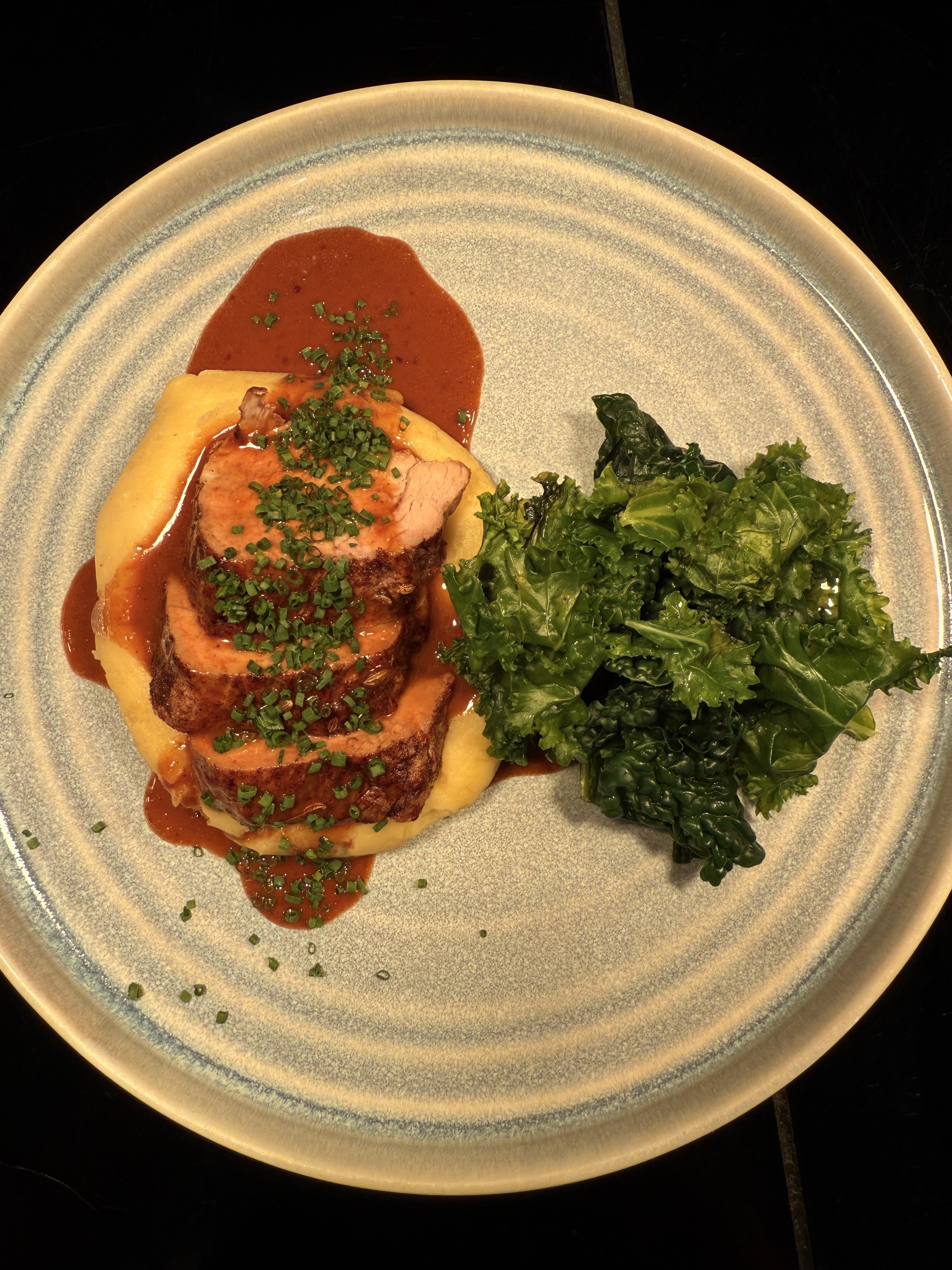 Plate with sliced roasted meat topped with herbs, served with mashed potatoes, gravy, and a side of kale salad.