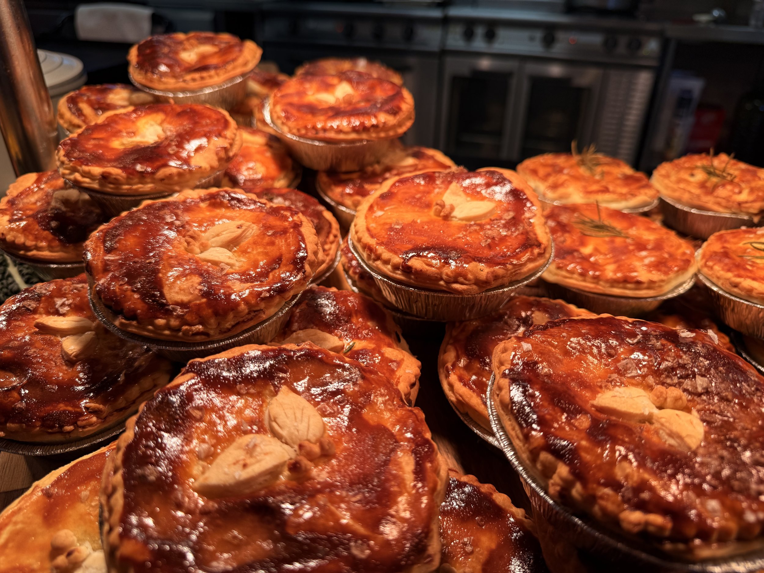 Several baked individual savory pies in aluminum cups with melted cheese and toppings, some garnished with nuts, displayed on a counter in a kitchen.