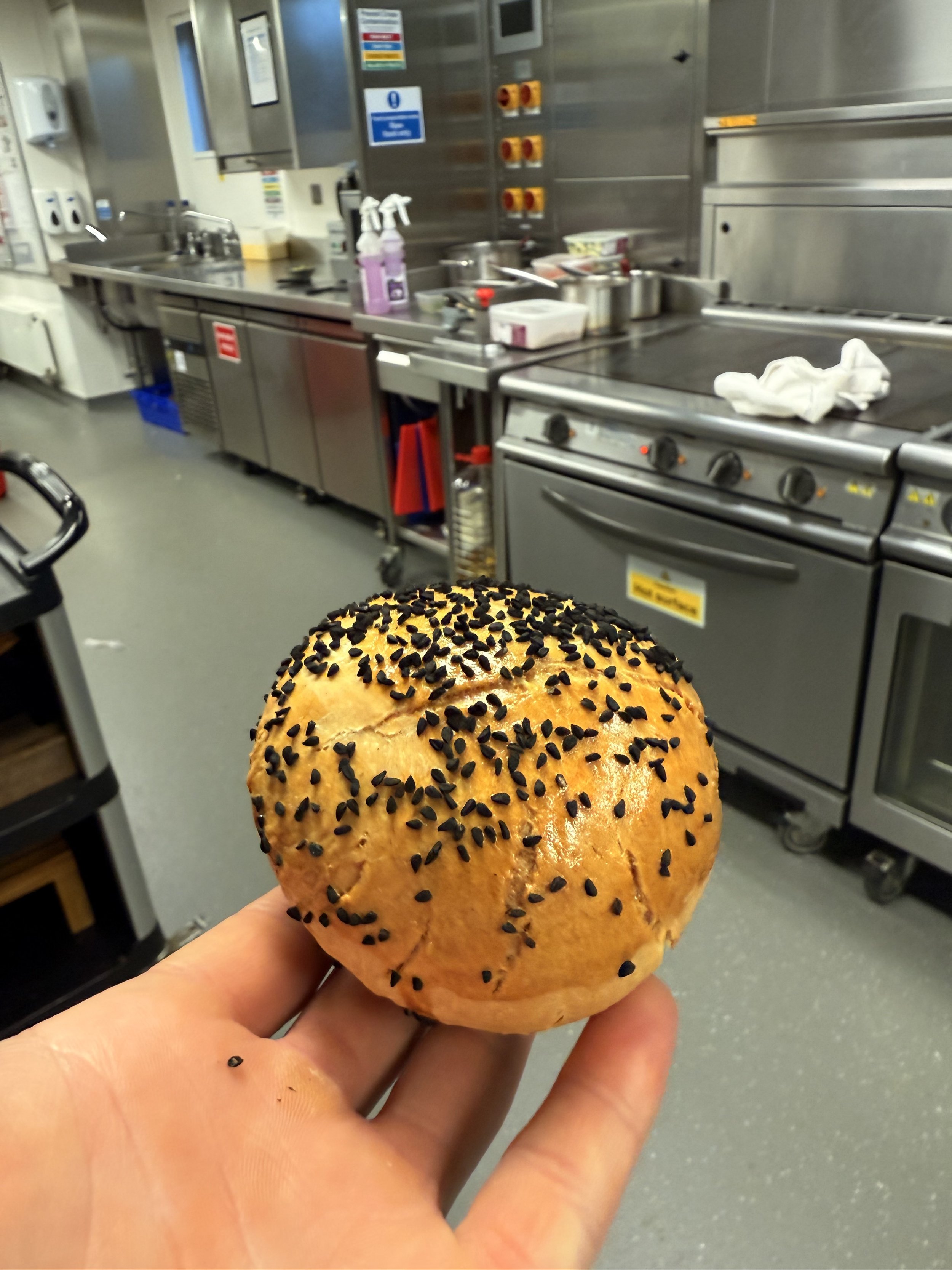 A hand holding a round bread roll topped with black sesame seeds in a commercial kitchen.
