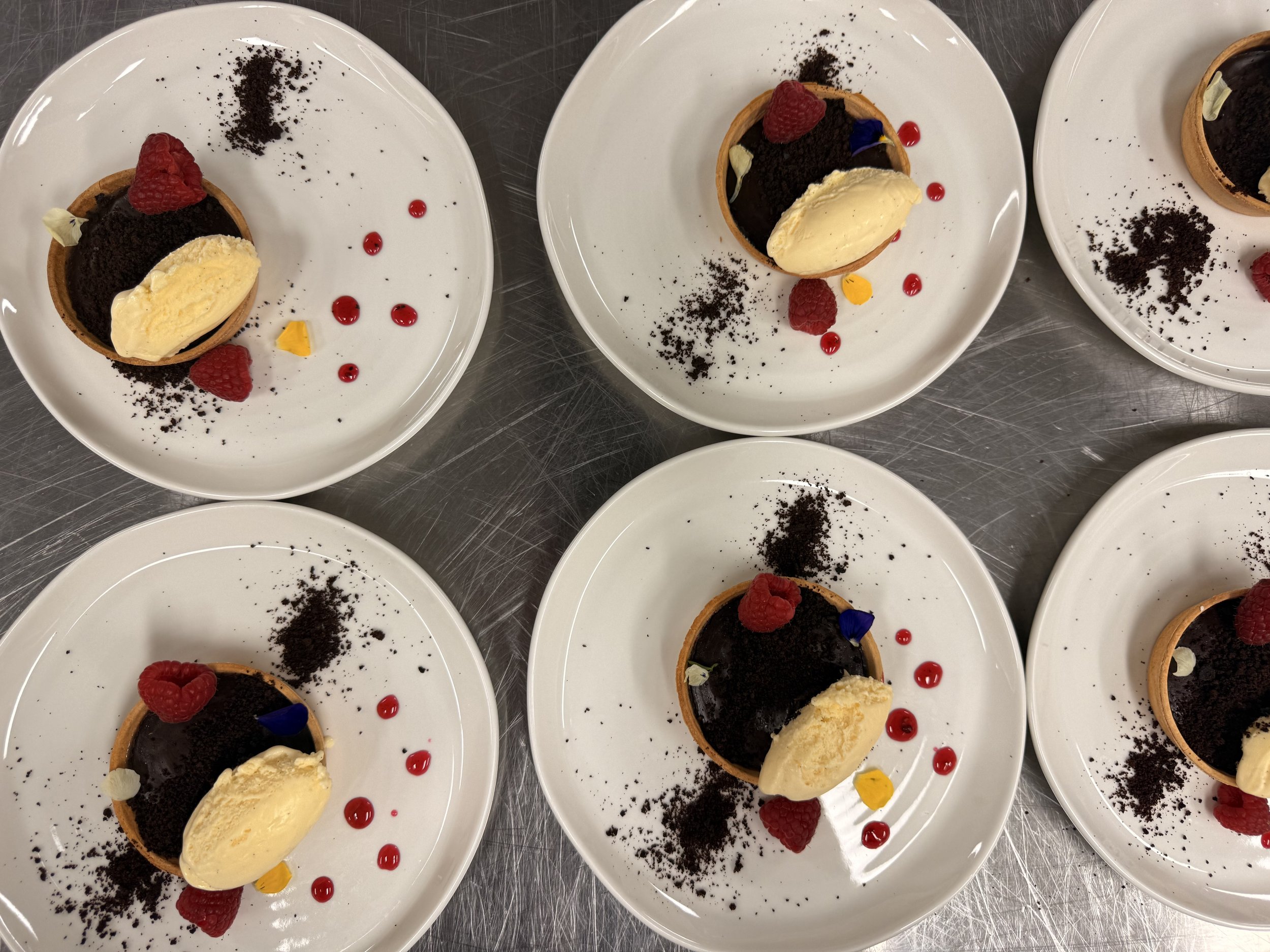 Multiple plates with small chocolate tarts garnished with raspberries, edible flowers, and red sauce dots, served with a scoop of vanilla ice cream.