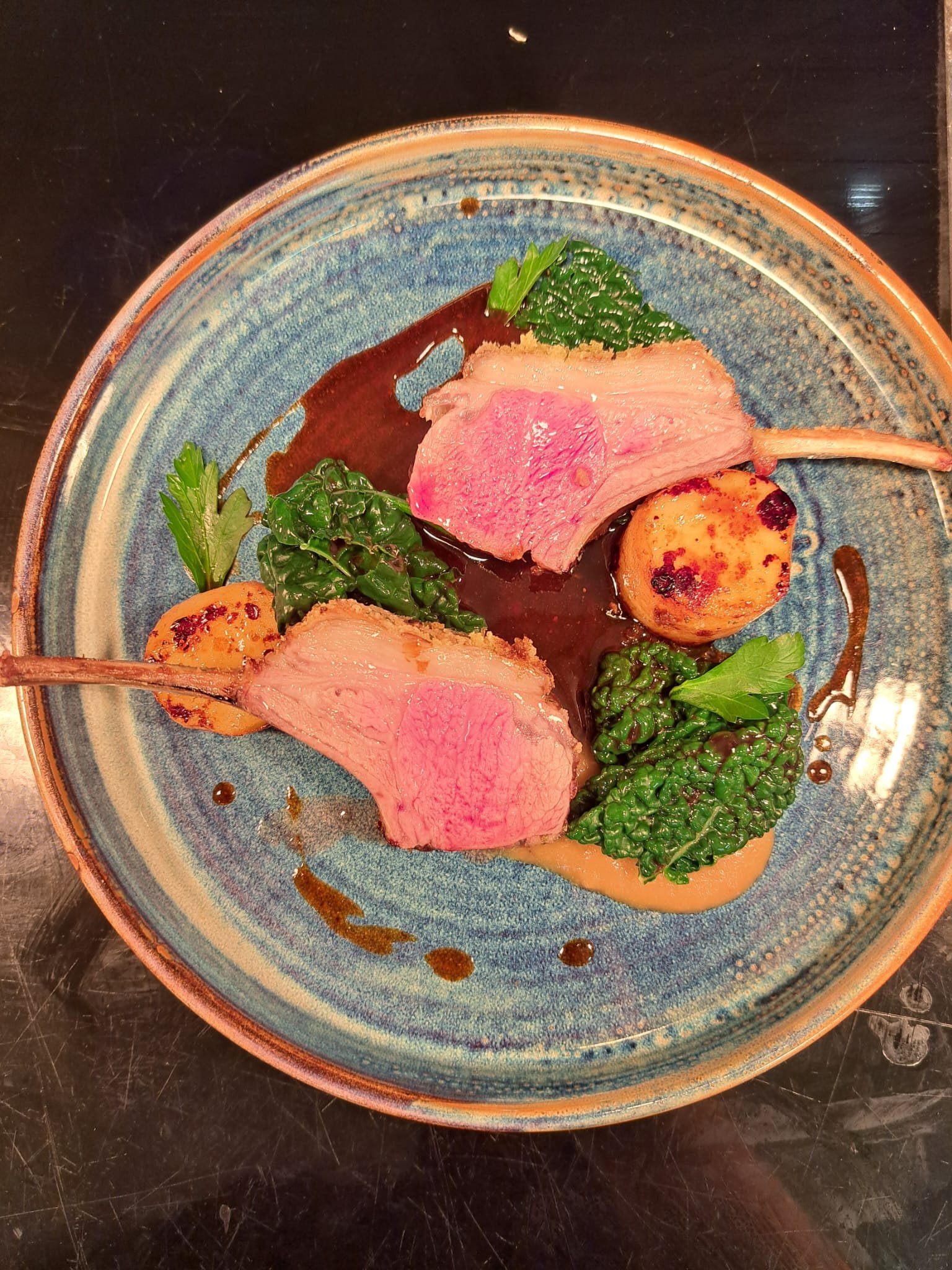 Two lamb chops on a blue ceramic plate, garnished with greens and accompanied by roasted potatoes, with sauce drizzled around.