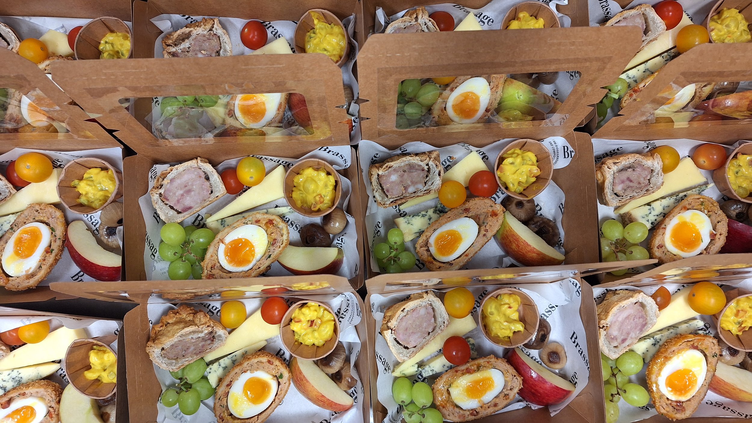Multiple small cardboard boxes filled with assorted savory snacks and fruits, including sliced egg cups, cheese, apple slices, green and red grapes, cherry tomatoes, and small bowls of mustard.