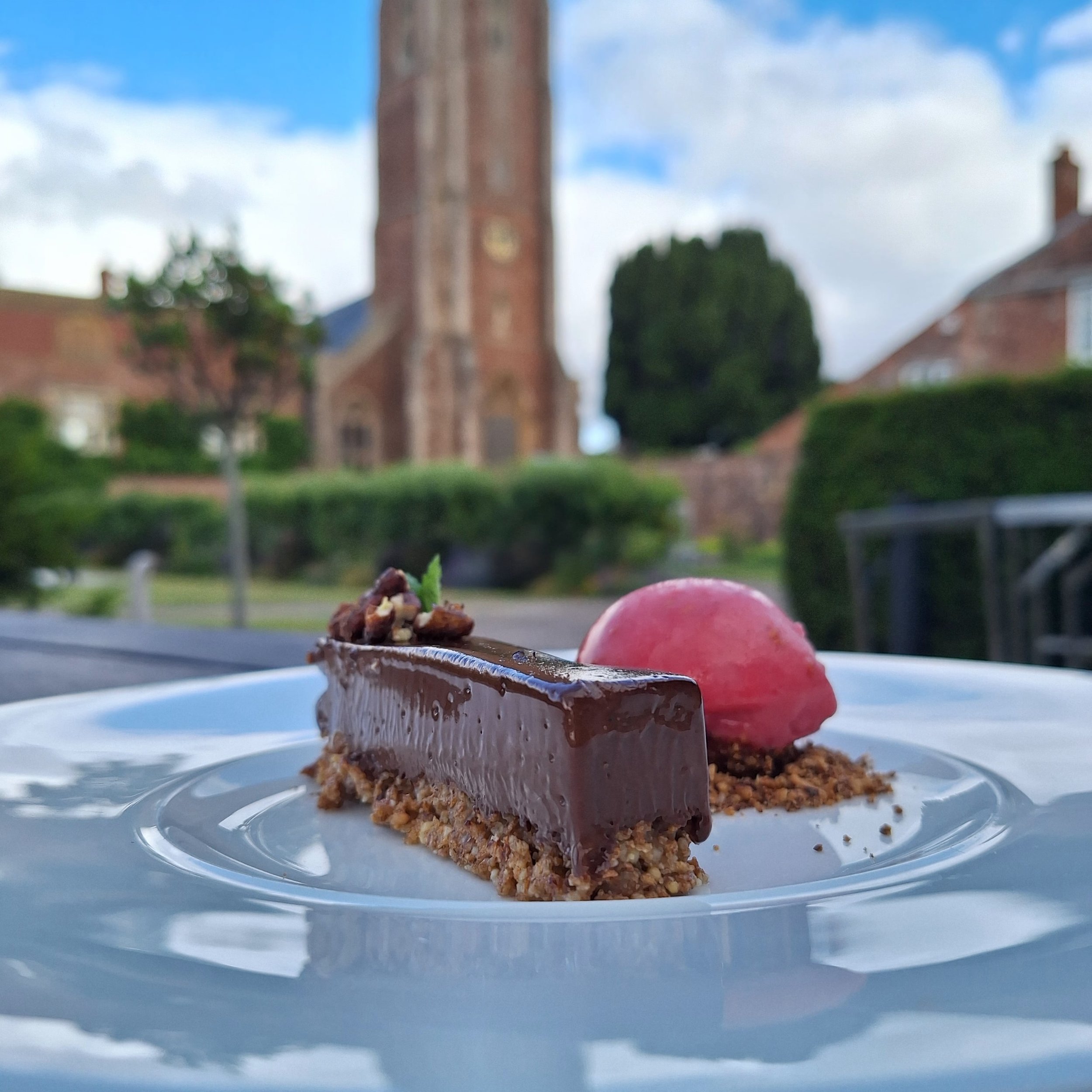 Chocolate dessert with a scoop of pink sorbet on a white plate, outdoors with a historic church and blue sky in the background.