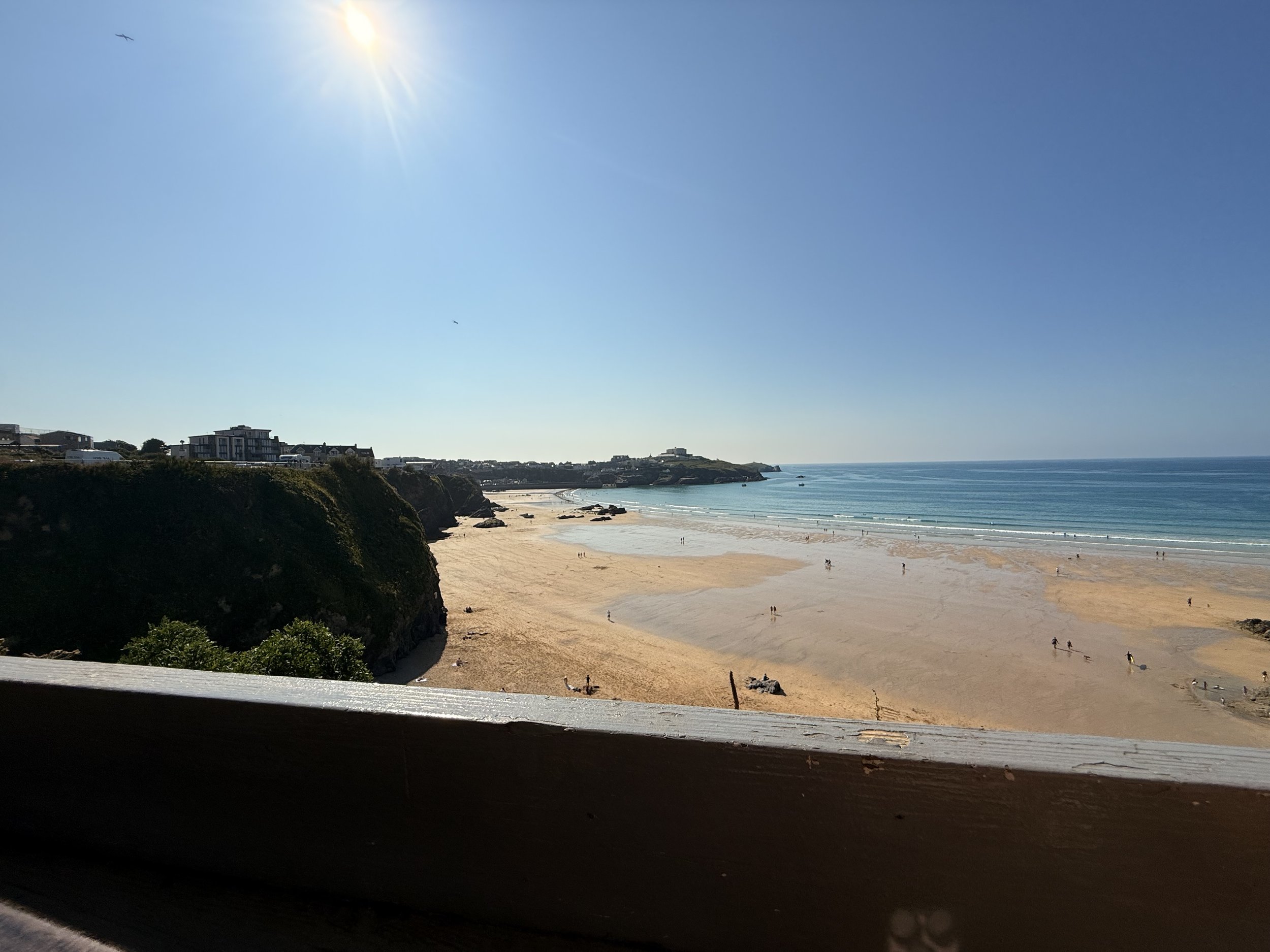 Sun shining over a sandy beach with cliffs on the left side, ocean waves, and a few people walking or relaxing on the beach under a clear blue sky.