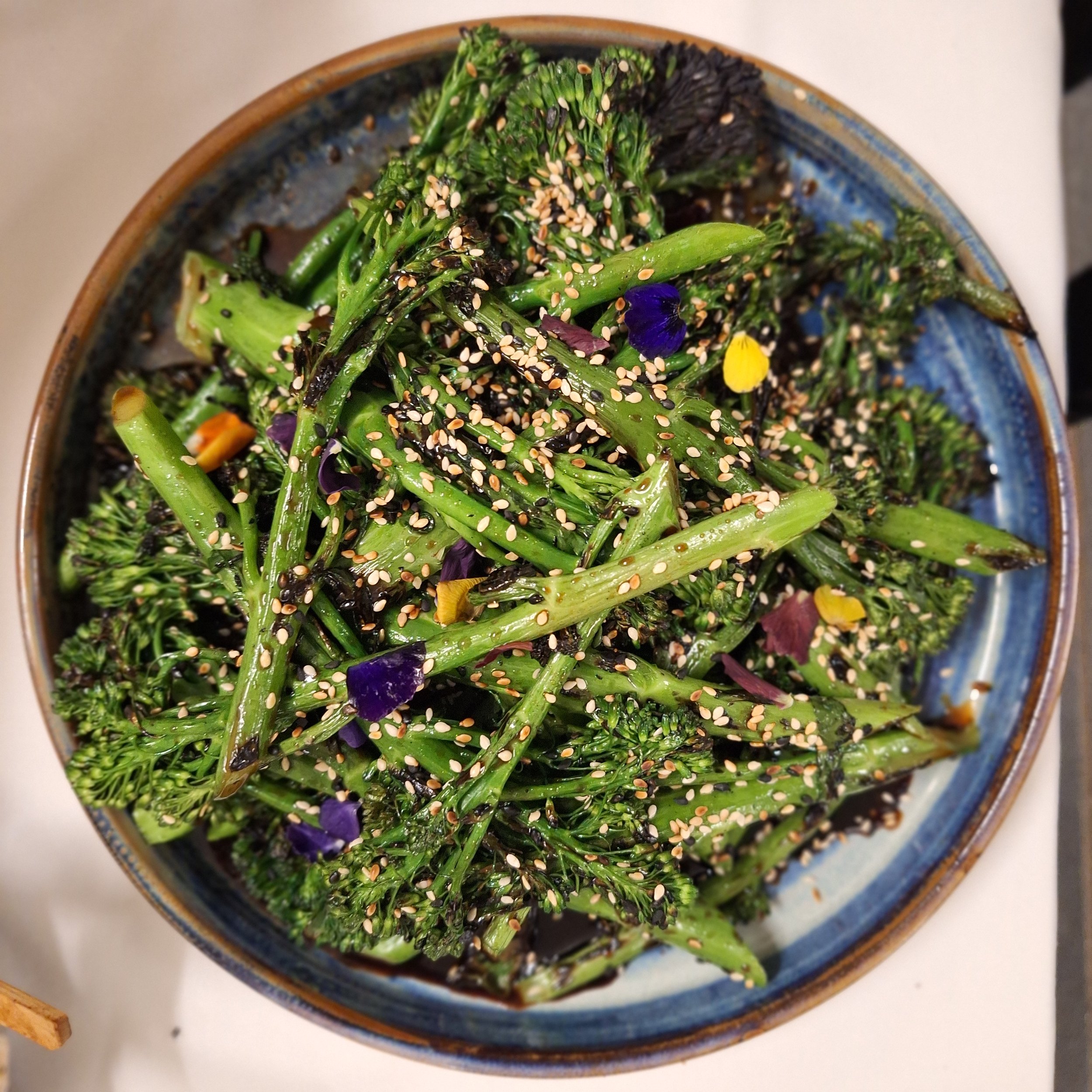 A bowl of cooked broccoli florets topped with sesame seeds and edible flowers.