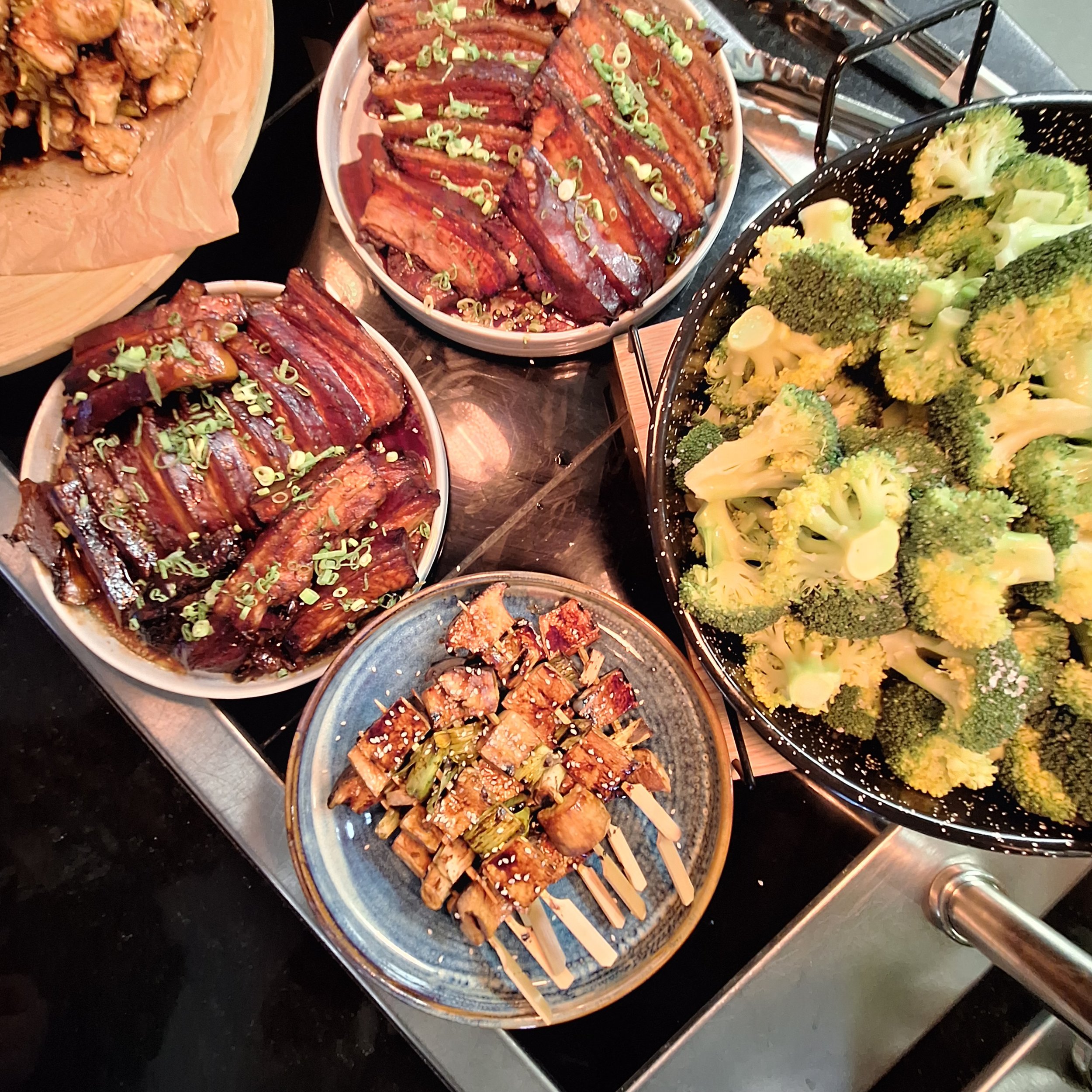 Assorted dishes of grilled and stir-fried vegetables including broccoli, eel, bacon-wrapped asparagus, and other Asian-inspired dishes on a buffet table.