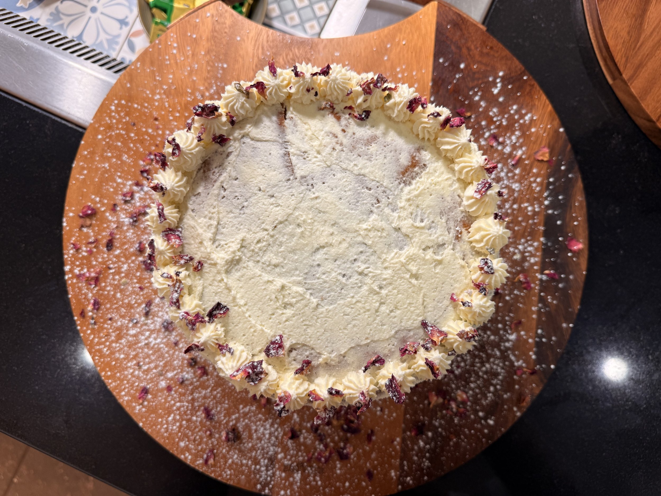 Round white cake with decorated border, dusted with powdered sugar, on a wooden cake board, with dried rose petals on top.