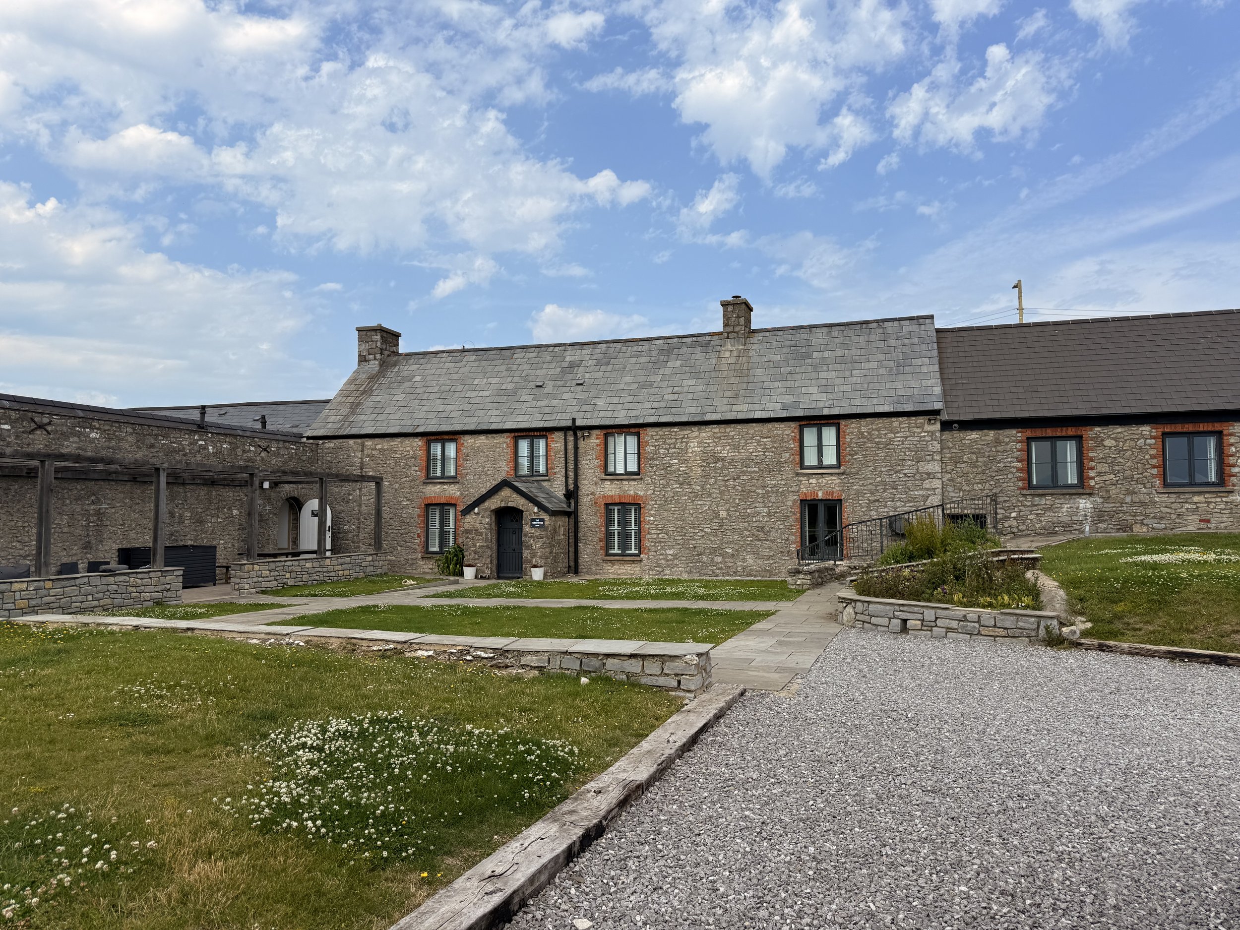A stone building with multiple windows and a central door, set against a partly cloudy sky, with a well-maintained lawn and paved pathways in the foreground.