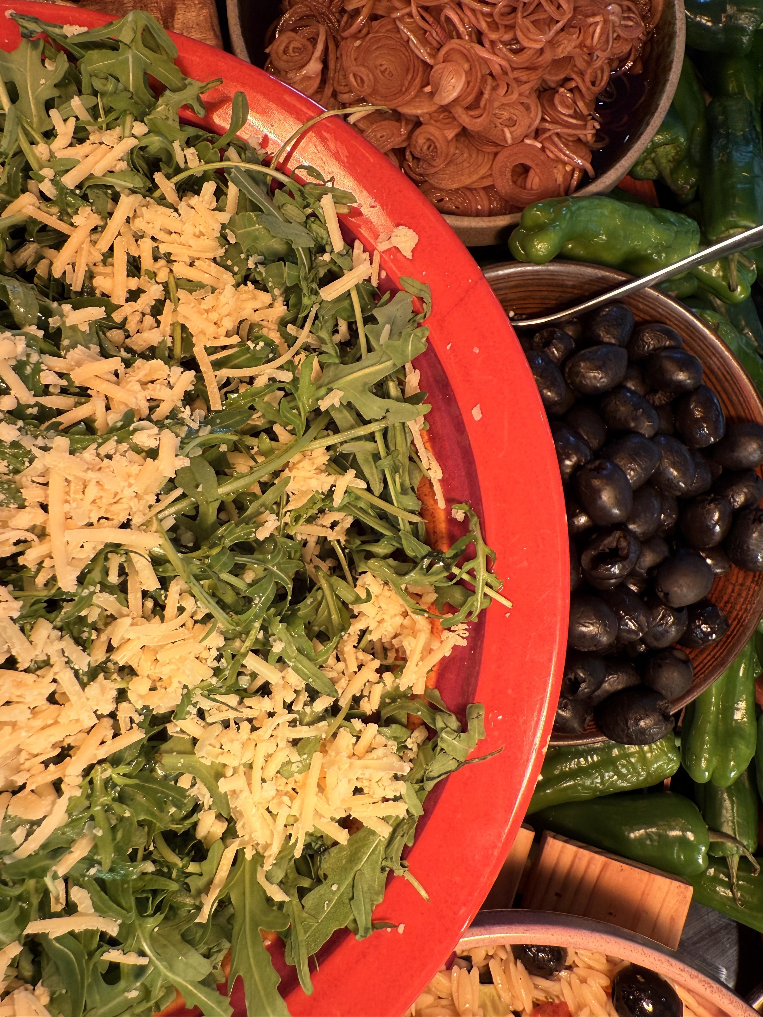 A close-up of a salad with arugula and shredded cheese in a red bowl, surrounded by bowls of black beans, chopped green peppers, pickled onions, and jalapeño peppers.