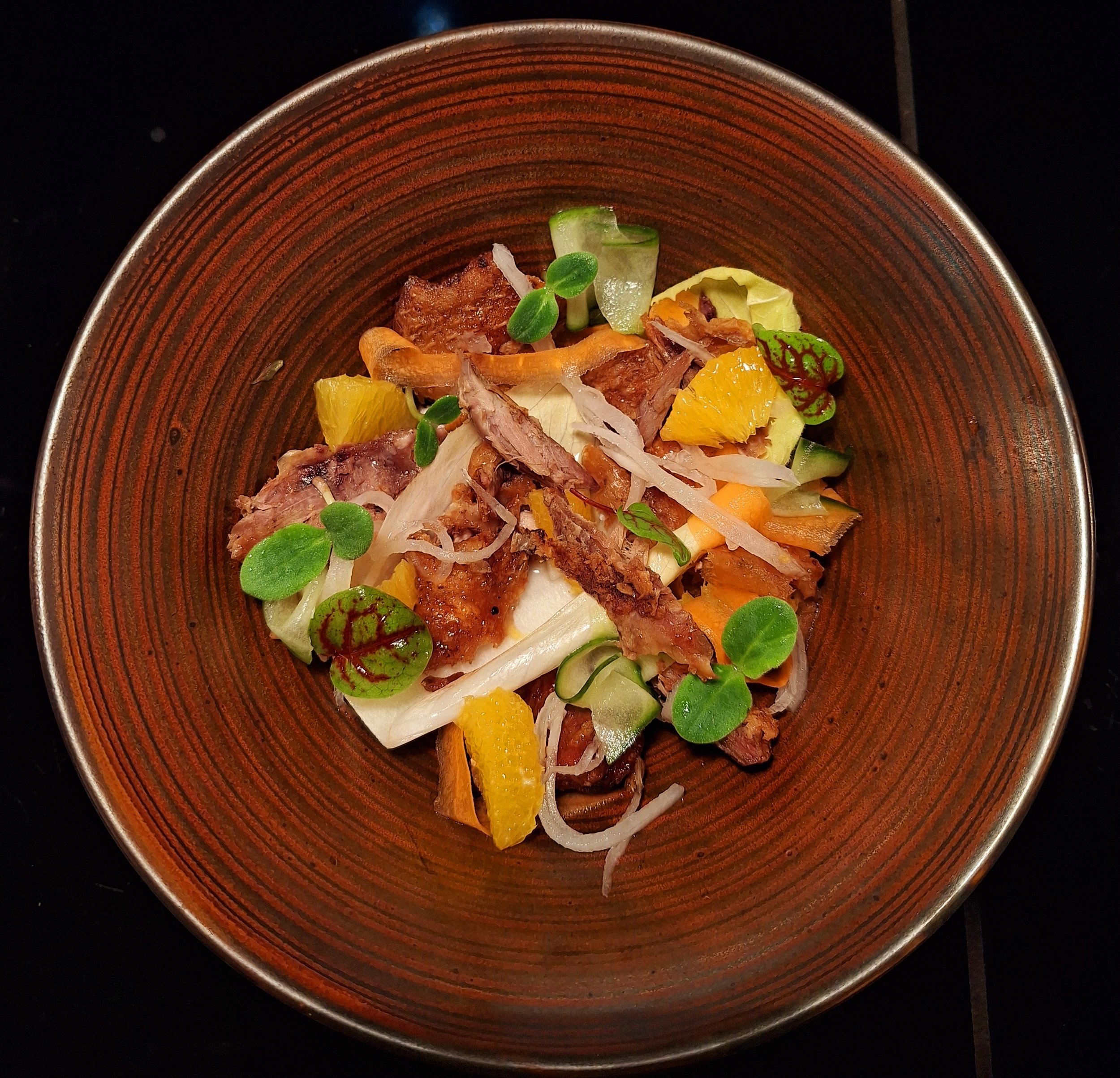 A plate of mixed salad with bacon, sliced vegetables, orange segments, and microgreens on a brown, textured plate.