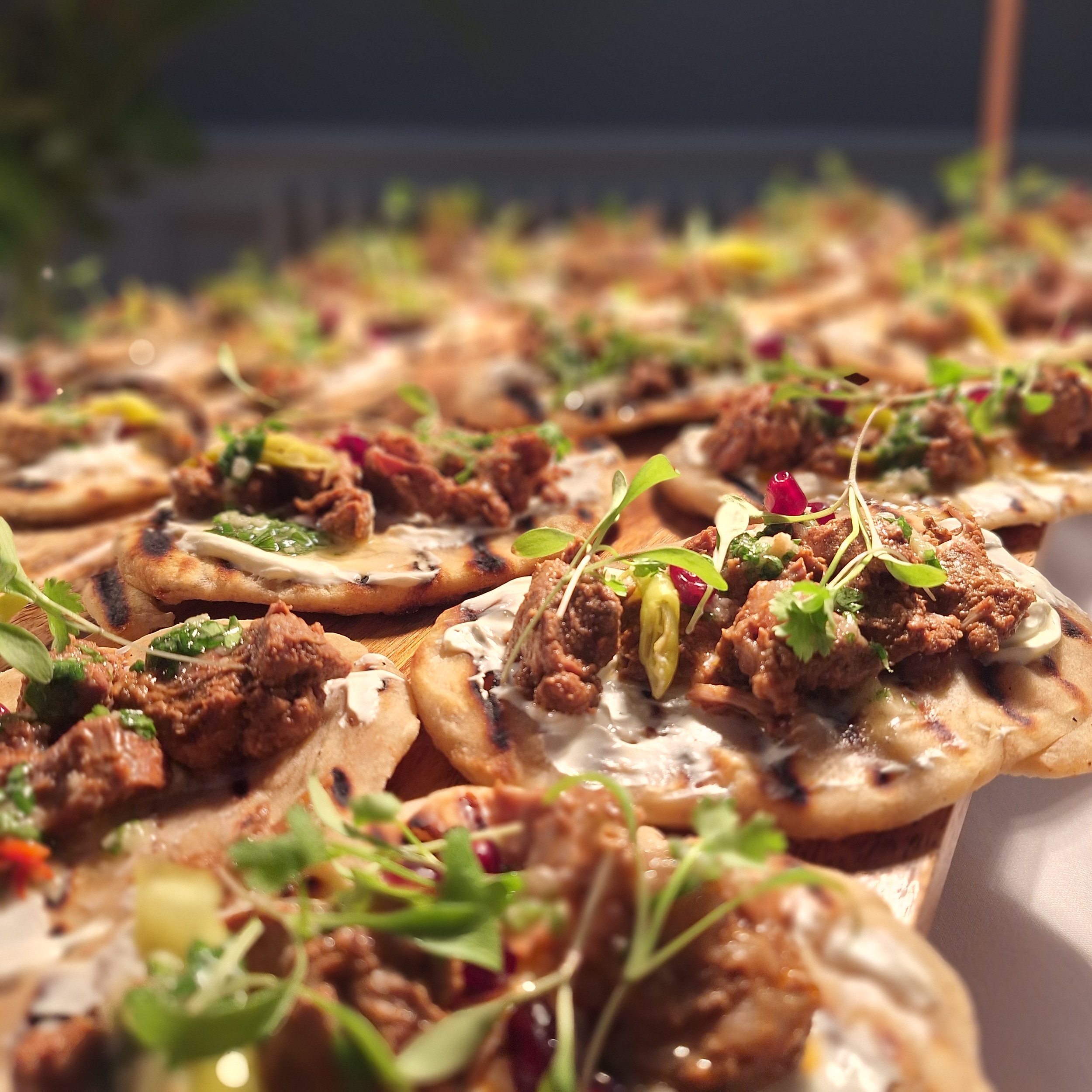 Close-up of small pizzas topped with beef, herbs, and microgreens on a wooden serving board.