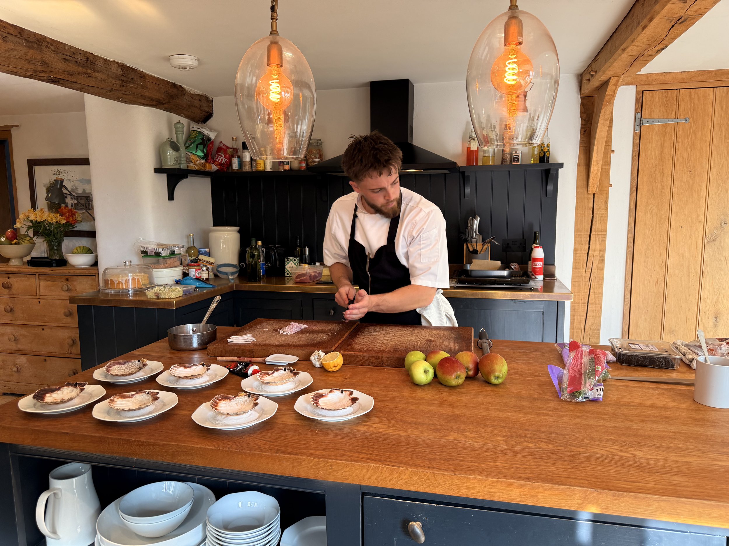 A man in a kitchen preparing food, with a wooden countertop, six plates with fish, apples, and various cooking supplies visible around him.