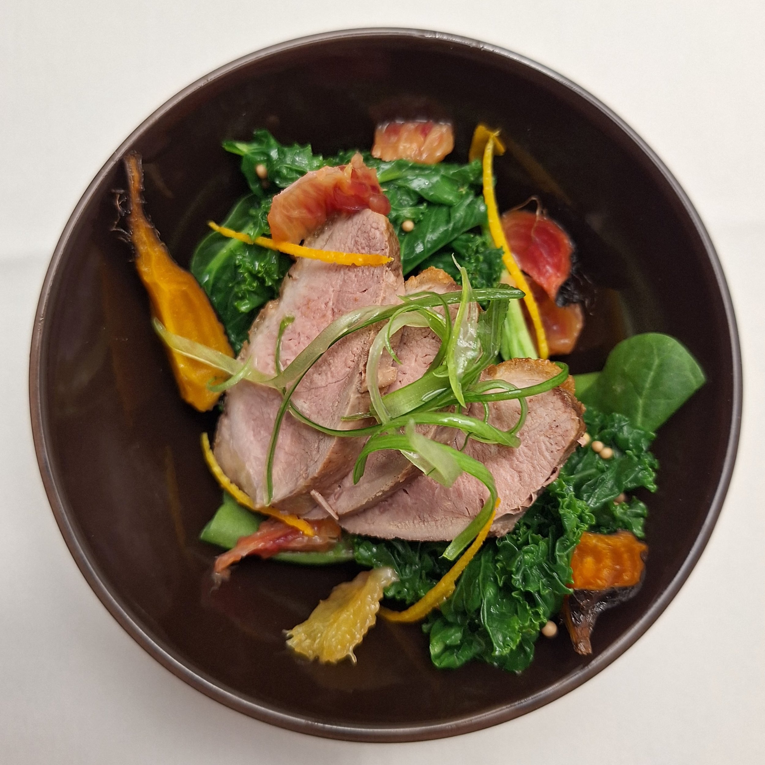 A bowl of gourmet salad with slices of cooked meat, kale, roasted vegetables, and garnished with fresh herbs.