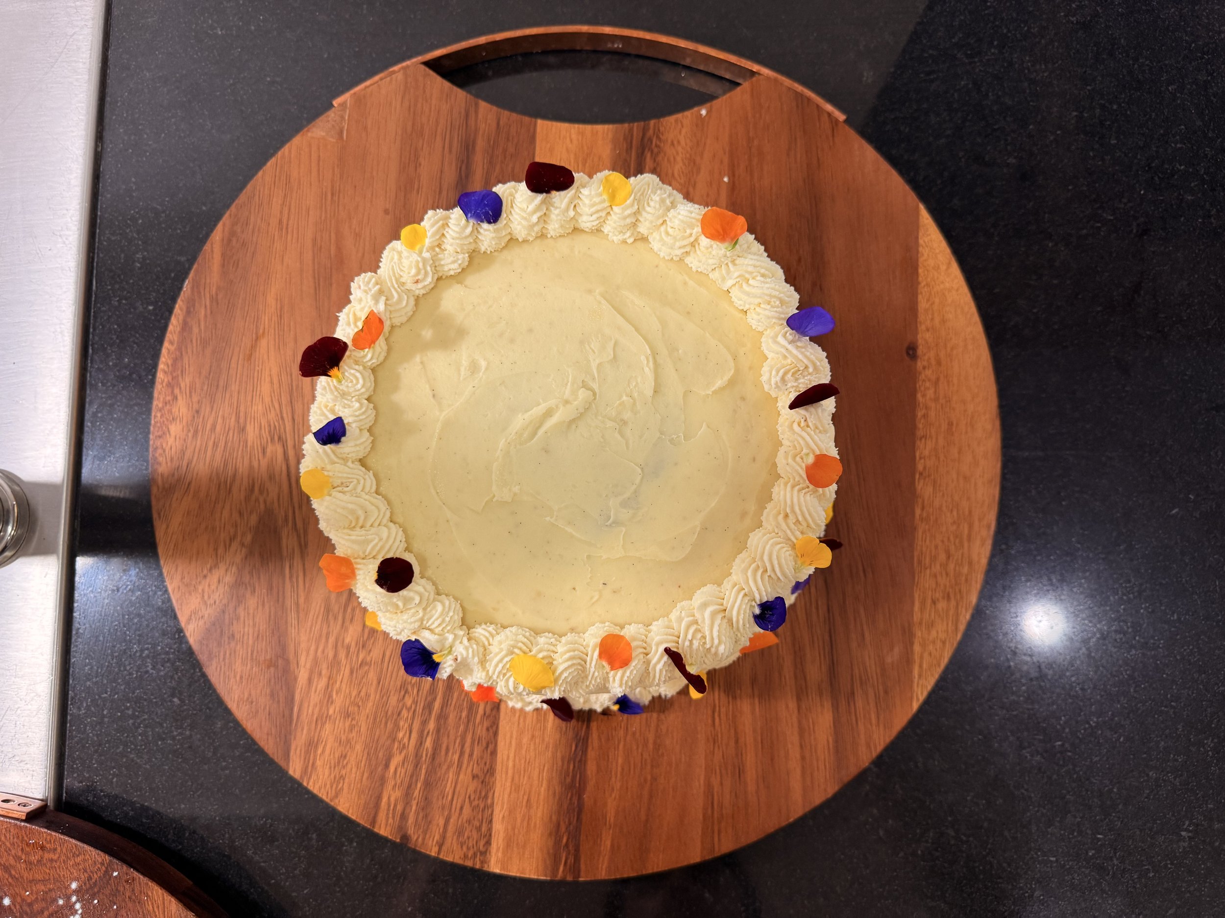 Round cake with white frosting border decorated with colorful edible flower petals, placed on a round wooden cake board.