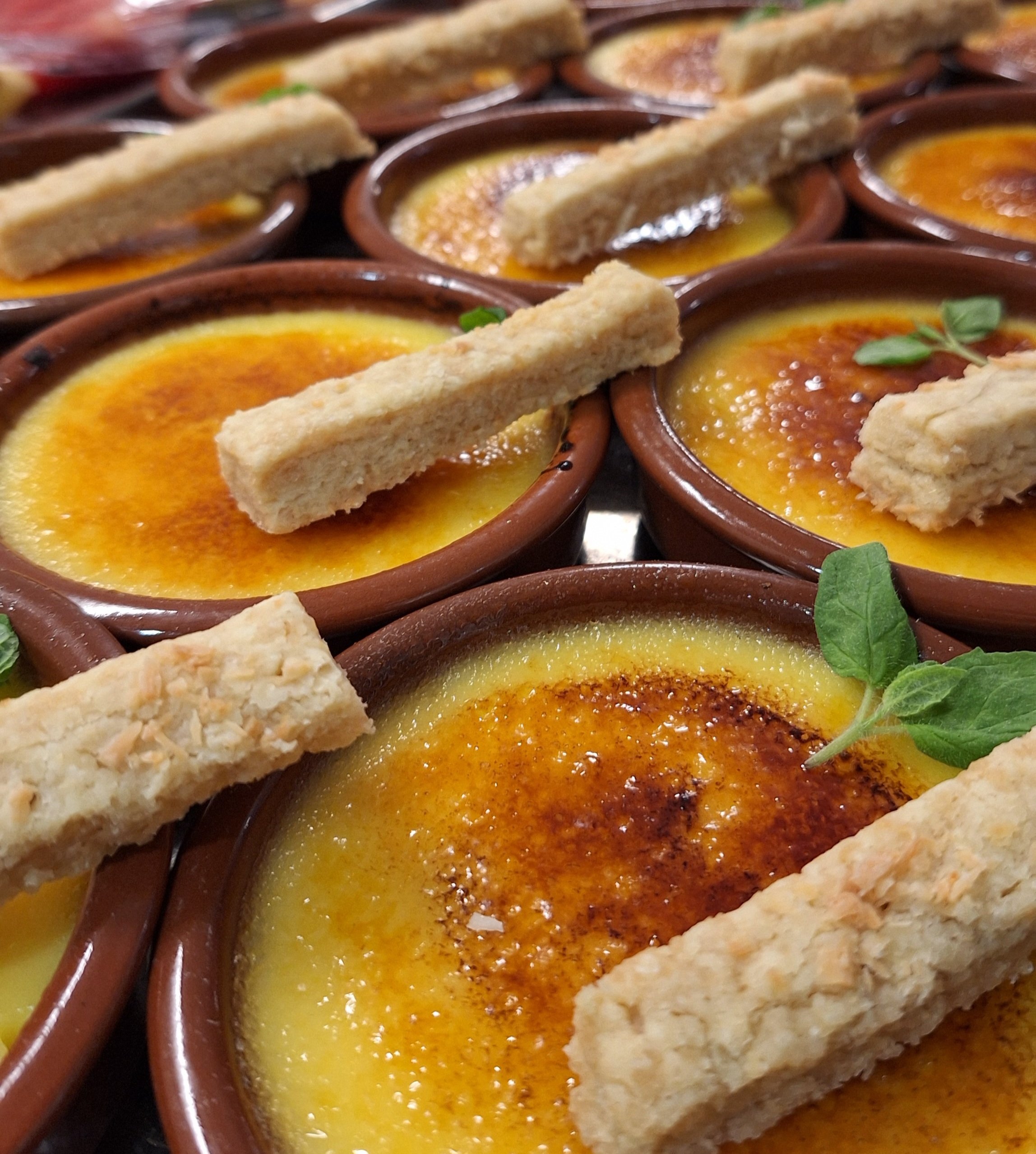 Close-up of multiple mini crème brûlées topped with a rectangular cookie and garnished with a small green leaf.